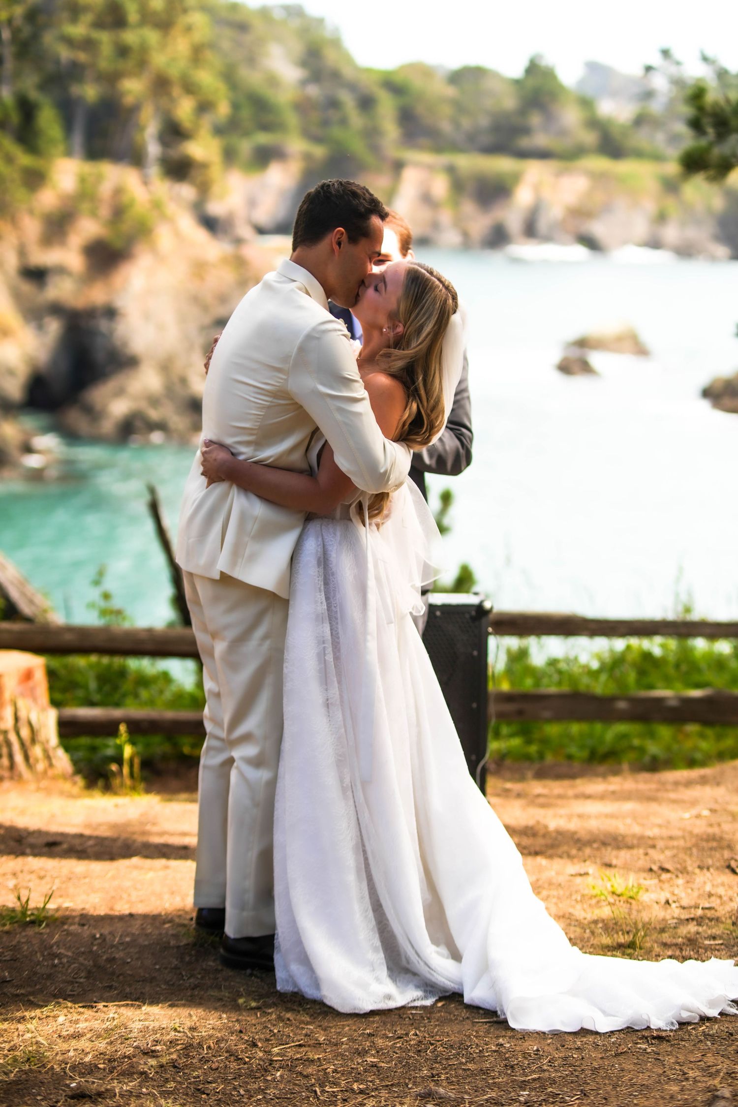 Bride and groom share a romantic kiss in white wedding attire with a scenic coastal ocean backdrop.