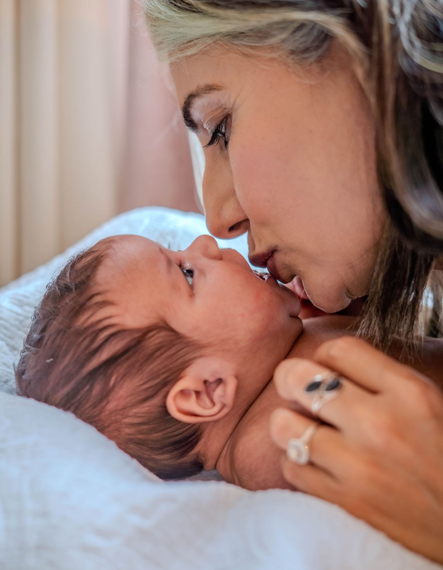 A loving mother gently kisses her newborn baby while cradling them, wearing a diamond engagement ring.