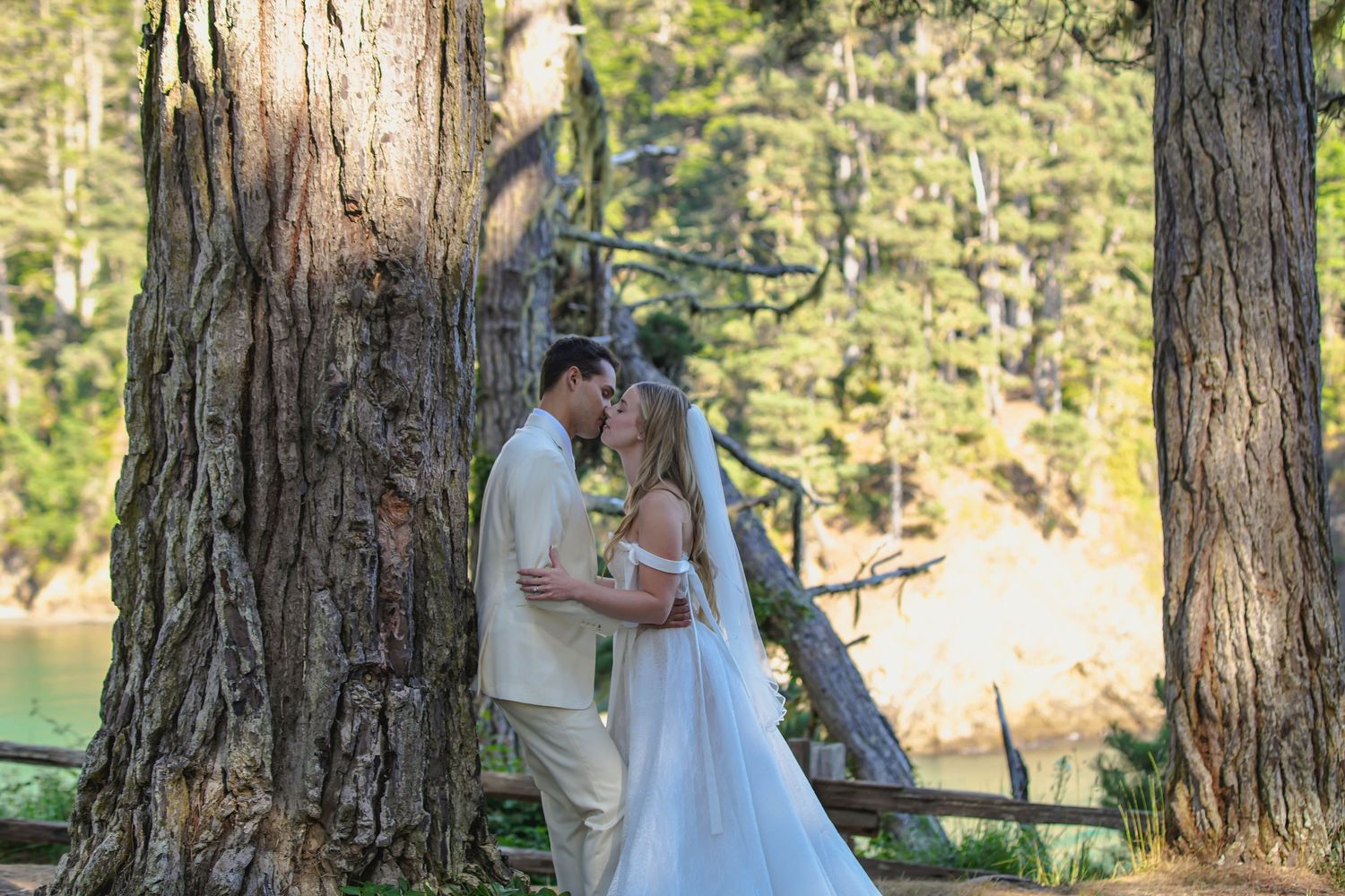 Bride and groom share a romantic moment leaning against a large tree trunk in a lush forest wedding setting.