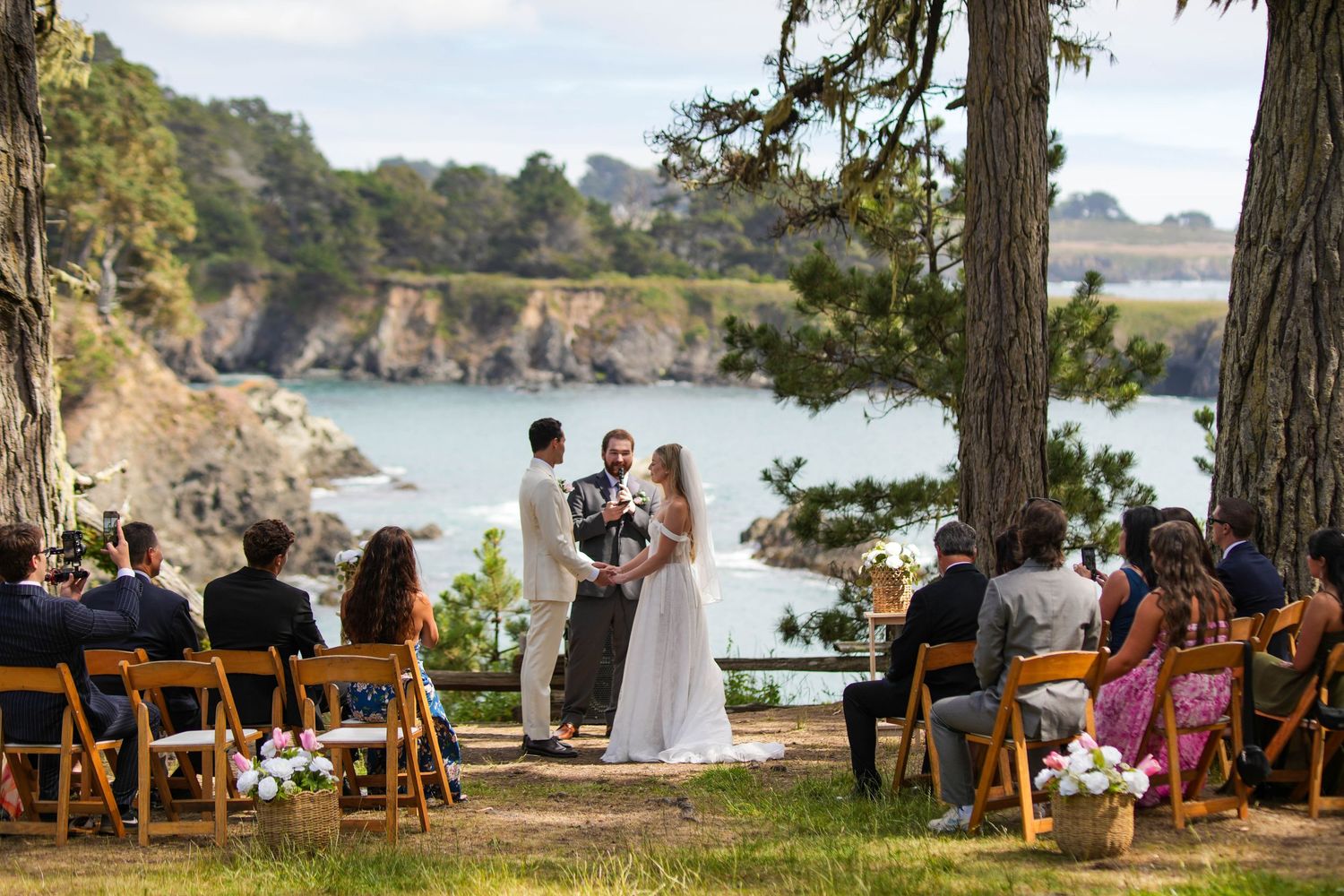 Outdoor wedding ceremony by ocean cliffs with couple at altar, guests seated on wooden chairs under pine trees.