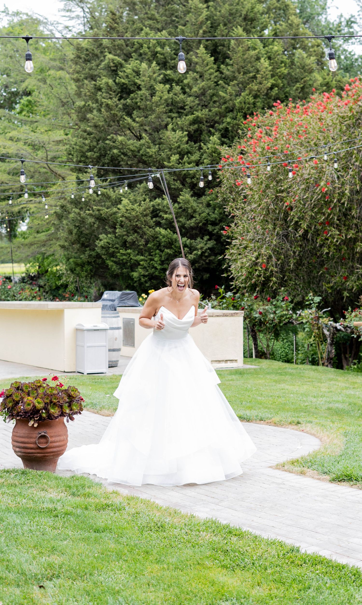White wedding dress displayed in an outdoor garden setting with red flowering bushes.