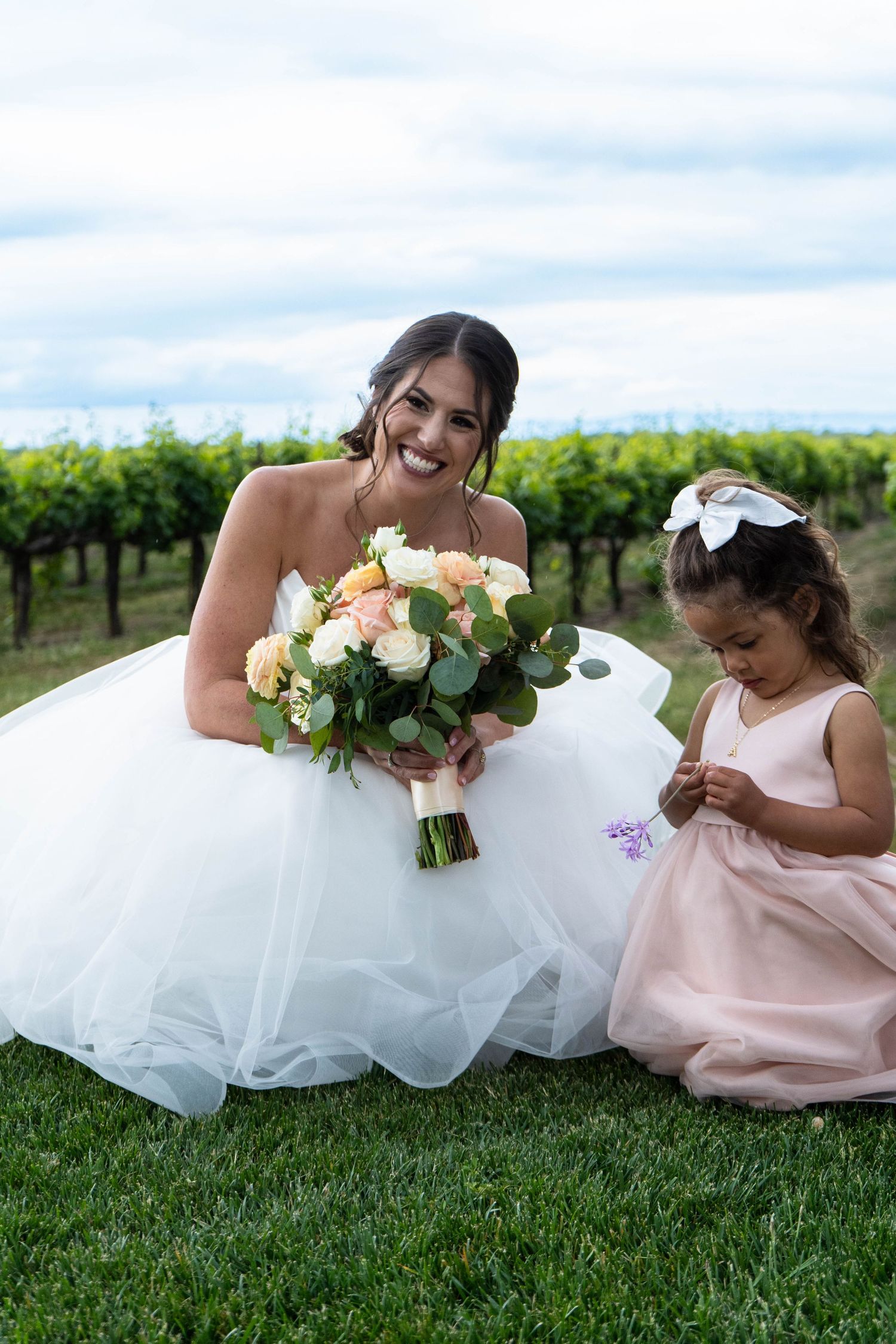 Bridesmaids in pink dresses and bride in white gown lay on grass holding bouquets at vineyard wedding.