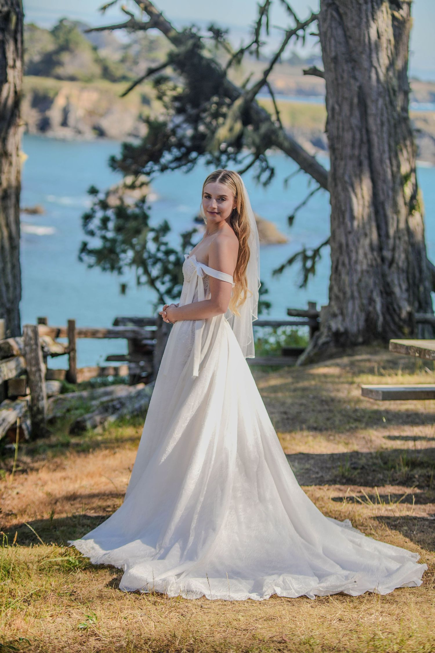 Bride in white off-shoulder gown with train poses outdoors near ocean and trees.