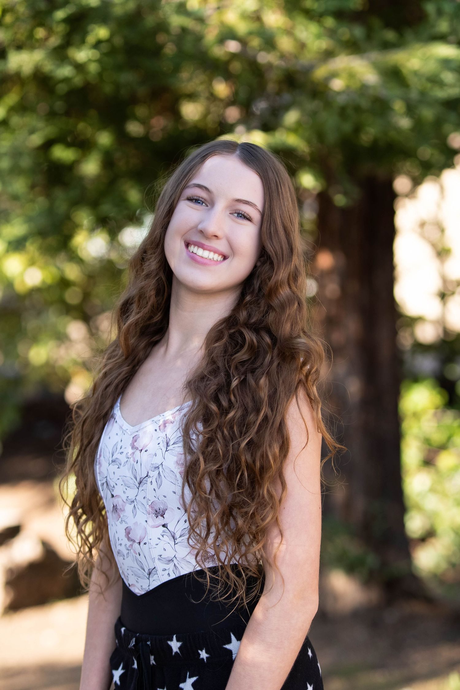 Young woman with long curly brown hair smiling outdoors, wearing a floral top and star-print skirt among green trees.