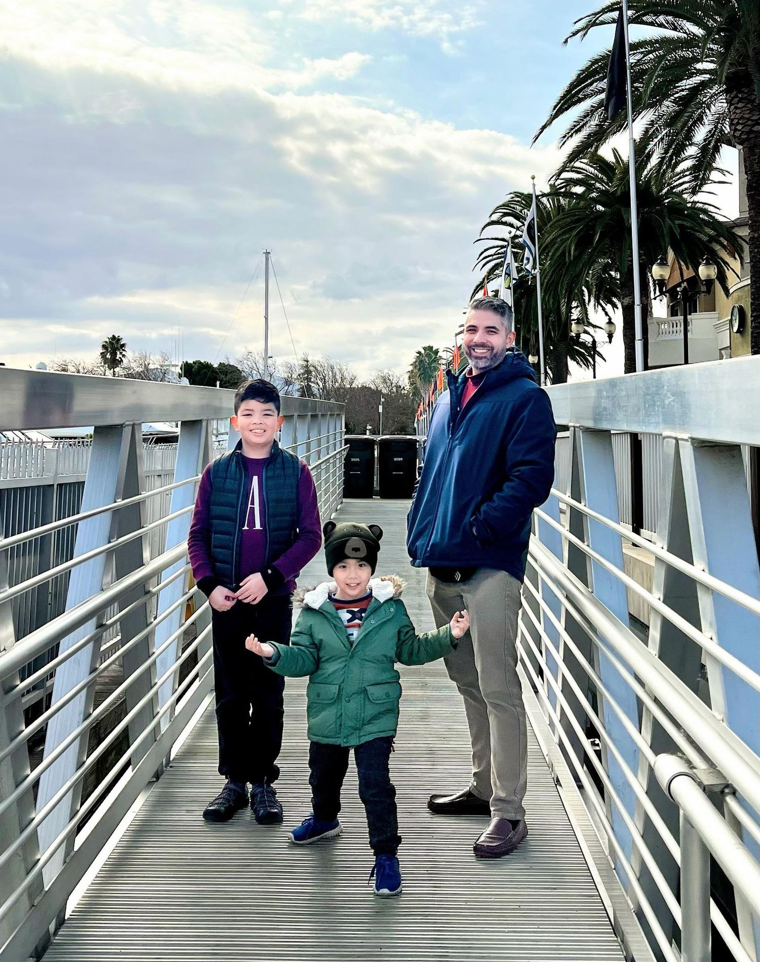 Group walking along a white-railed boardwalk near palm trees on an overcast day.