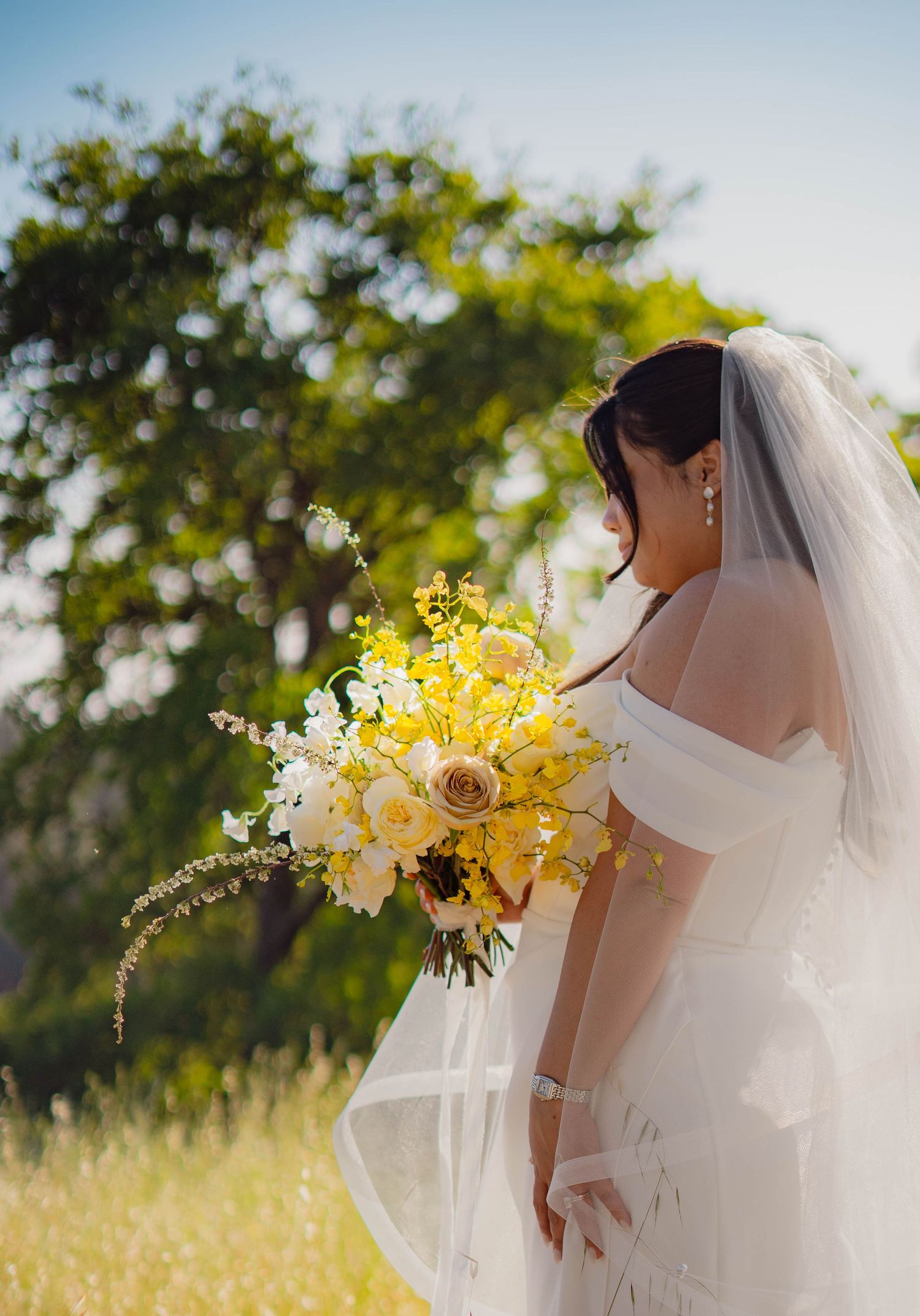 Bride in off-shoulder gown and veil holds yellow and white bouquet outdoors with lush green trees in background.