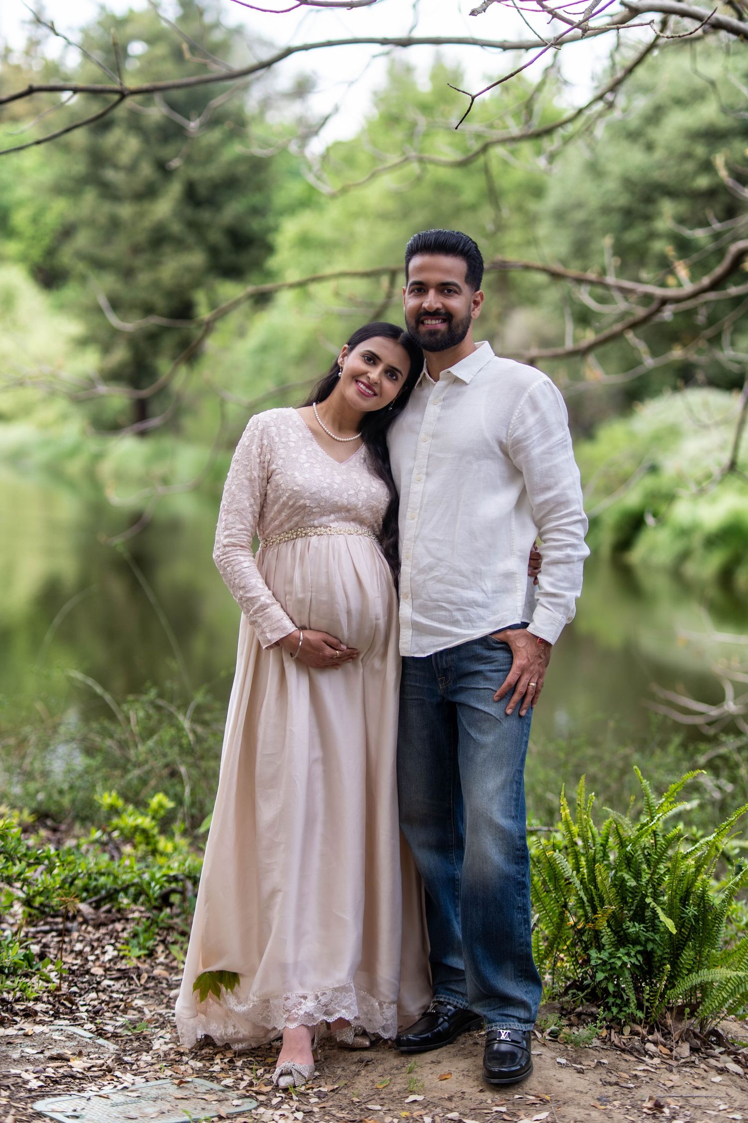 Pregnant couple posing outdoors near a pond surrounded by lush greenery, woman in elegant gown cradling baby bump.