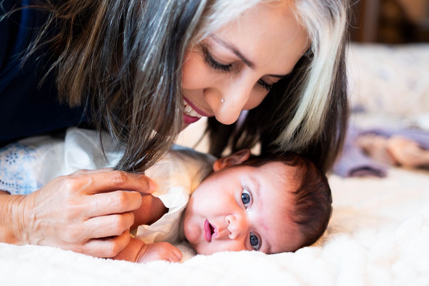 Grandmother with gray hair smiling and bonding with her newborn baby lying on a soft white blanket.