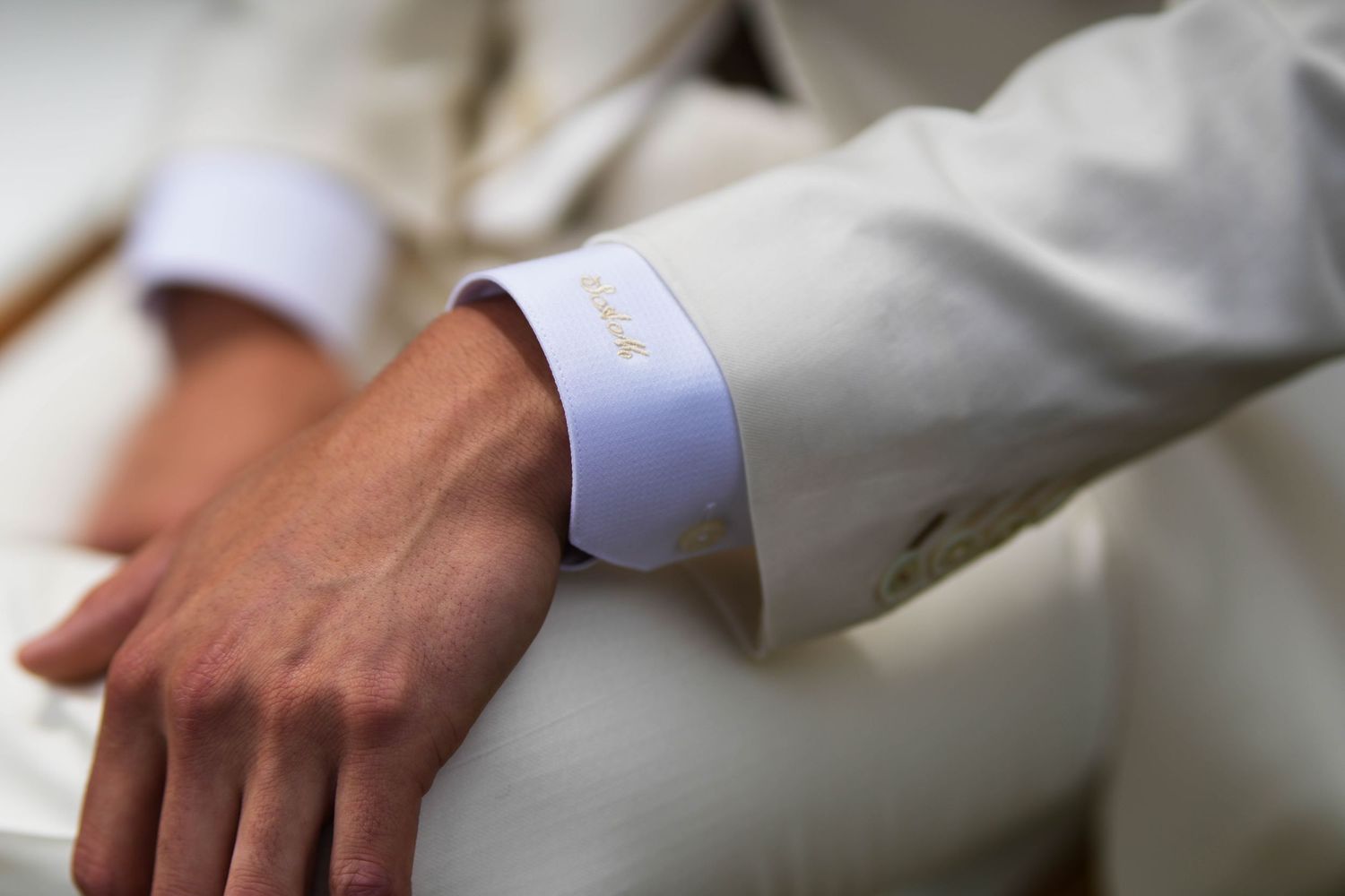 Close-up of a man's wrist in a white suit with an elegant monogrammed French cuff shirt detail.