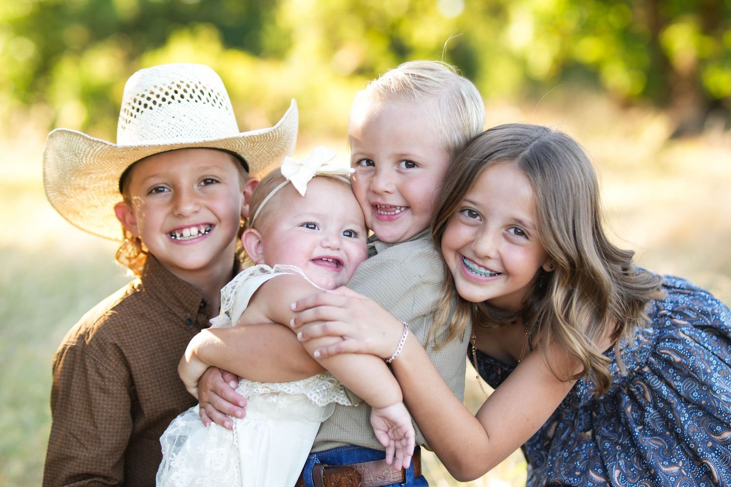 Four happy children hugging outdoors, oldest boy wearing cowboy hat, smiling siblings posing together in sunny park setting.