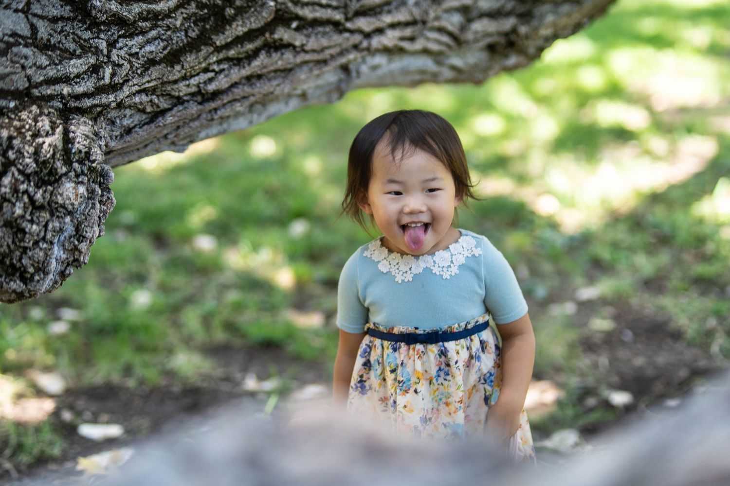 A child in a blue and floral dress laughs while playing near a tree.