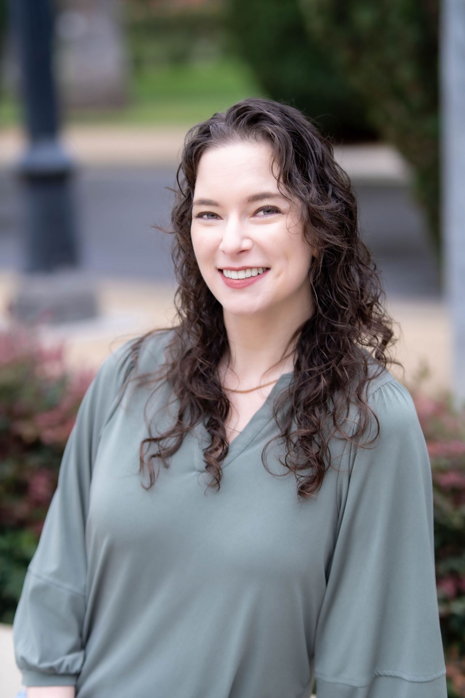 A person in a sage green top smiles warmly while posing outdoors in a natural setting.