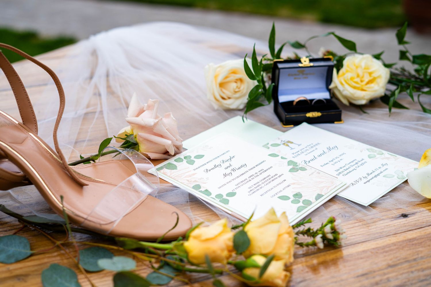 Wedding flat lay with nude heels, yellow roses, ring box, and elegant invitation cards on rustic wood.