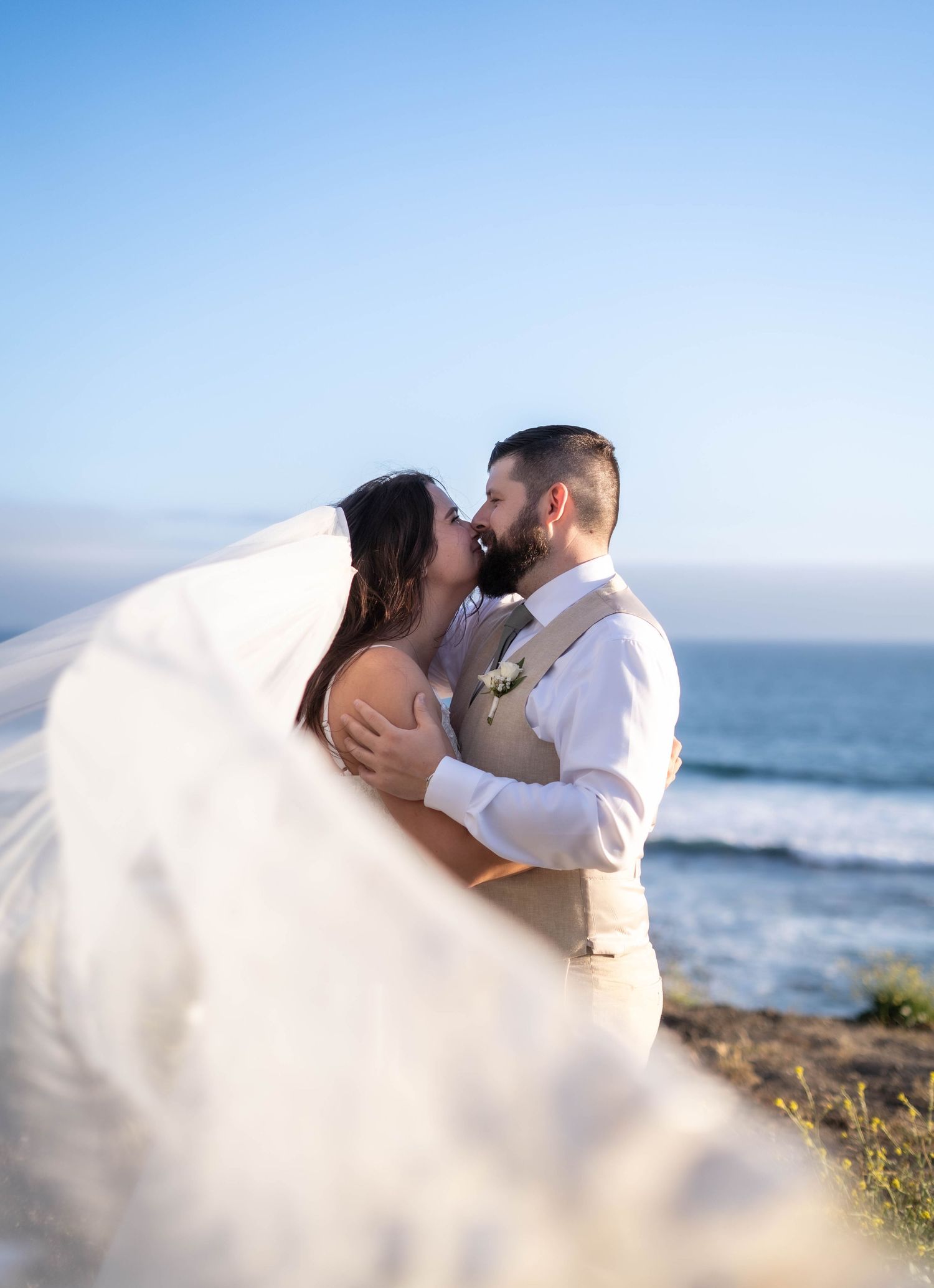 A bride and groom share a romantic kiss on a coastal cliff, her veil billowing in the ocean breeze.