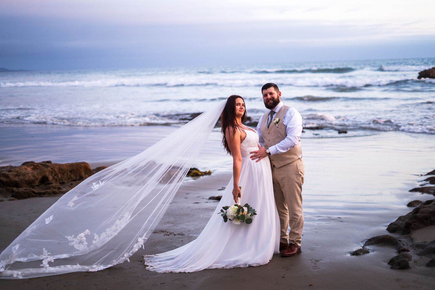 Bride and groom pose on a sandy beach at sunset, her long veil flowing in the wind with ocean waves behind them.