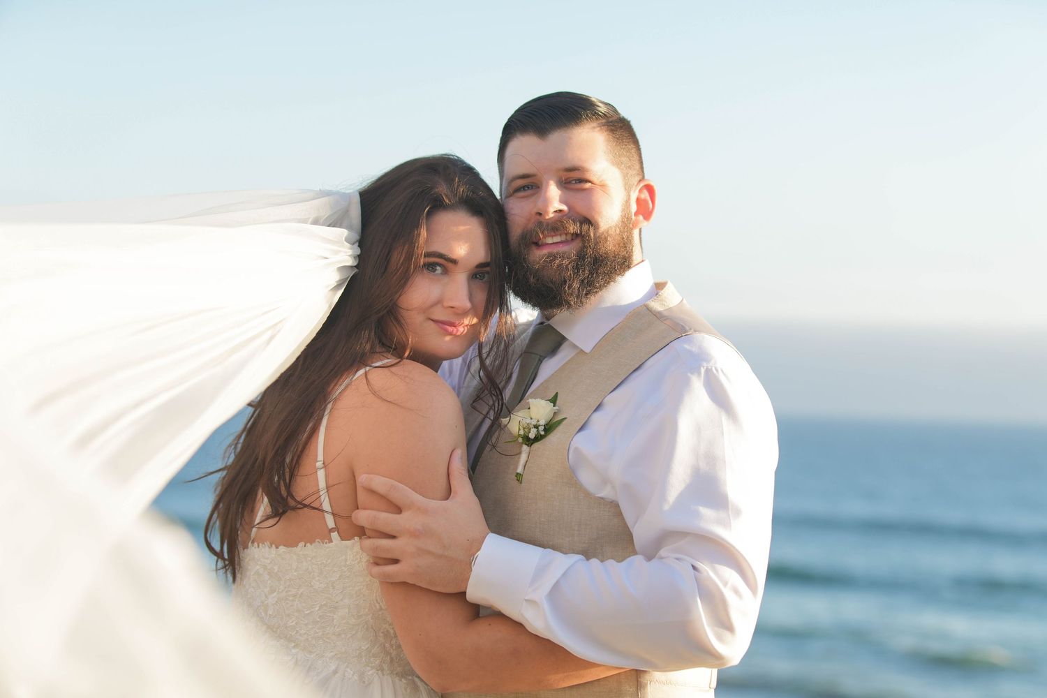 Bride and groom embrace on beach, veil flowing in wind, ocean backdrop at golden hour.