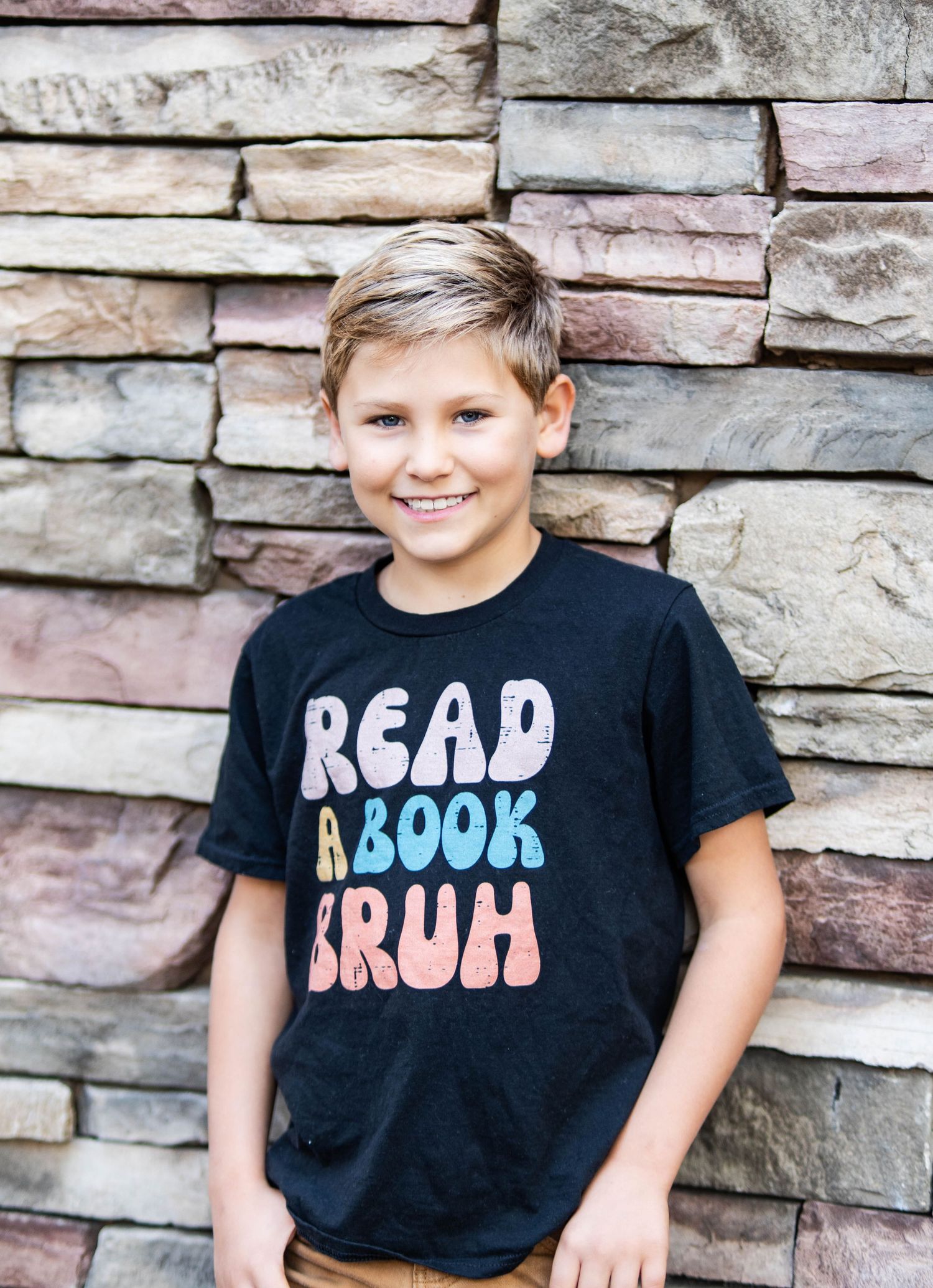 Boy smiling in black 'Read A Book Bruh' t-shirt, standing against a stacked stone wall background.