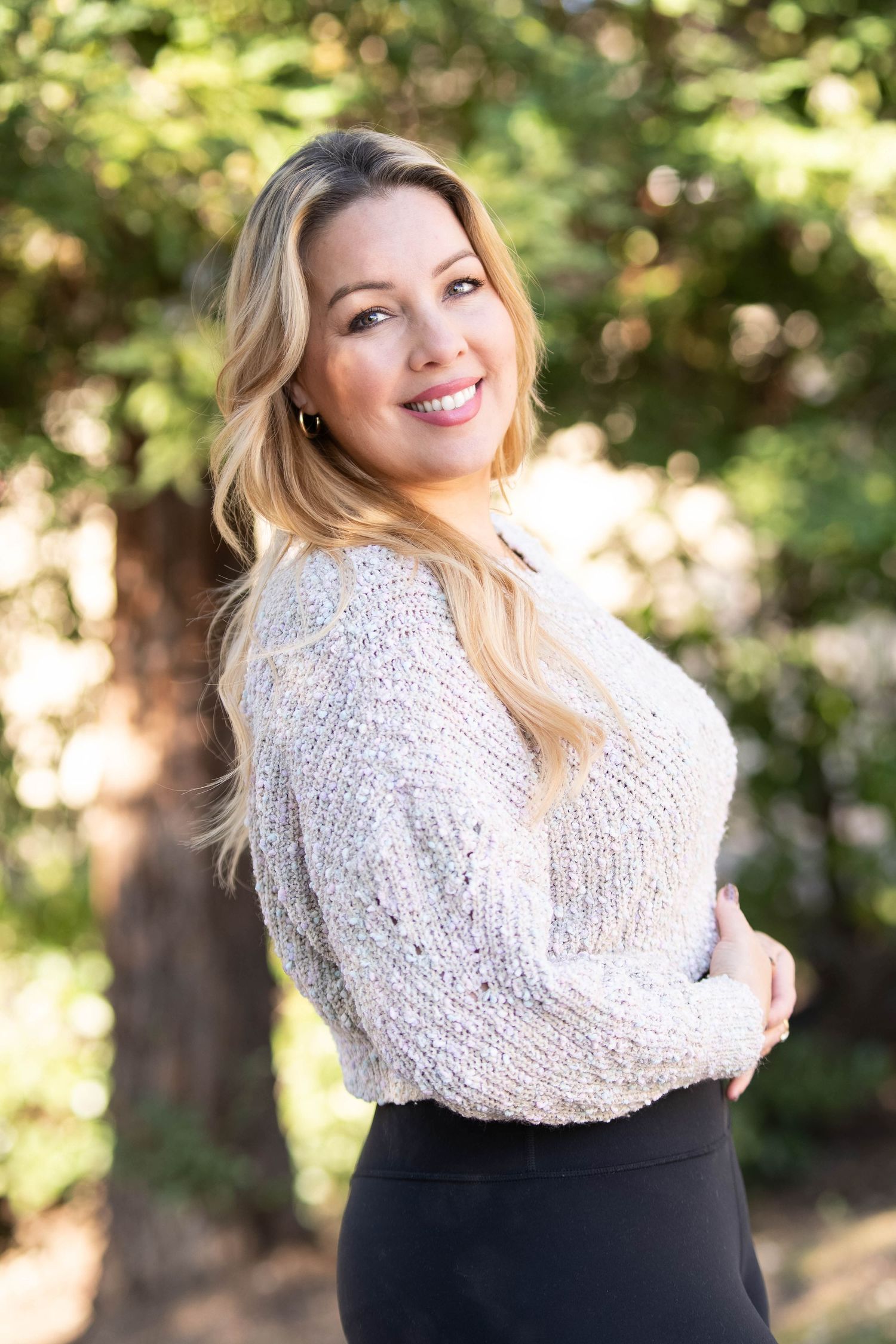 Smiling blonde woman in a white knit sweater and black skirt poses outdoors with arms crossed, trees in background.