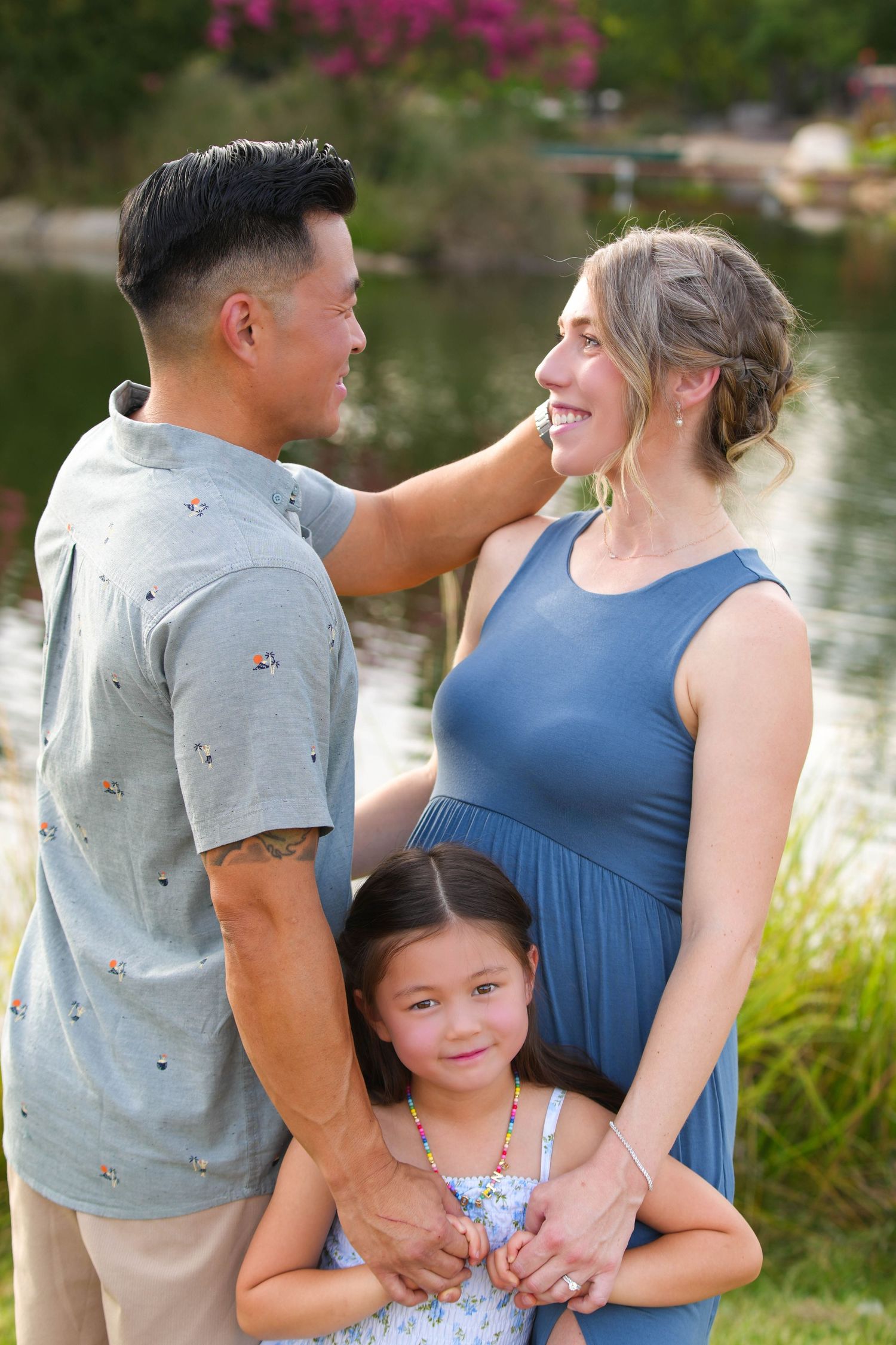 A happy family of three poses outdoors near a pond, with parents gazing at each other and daughter standing between them.