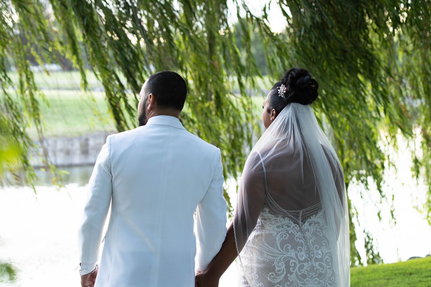 Wedding couple walking together under cascading willow branches near water.