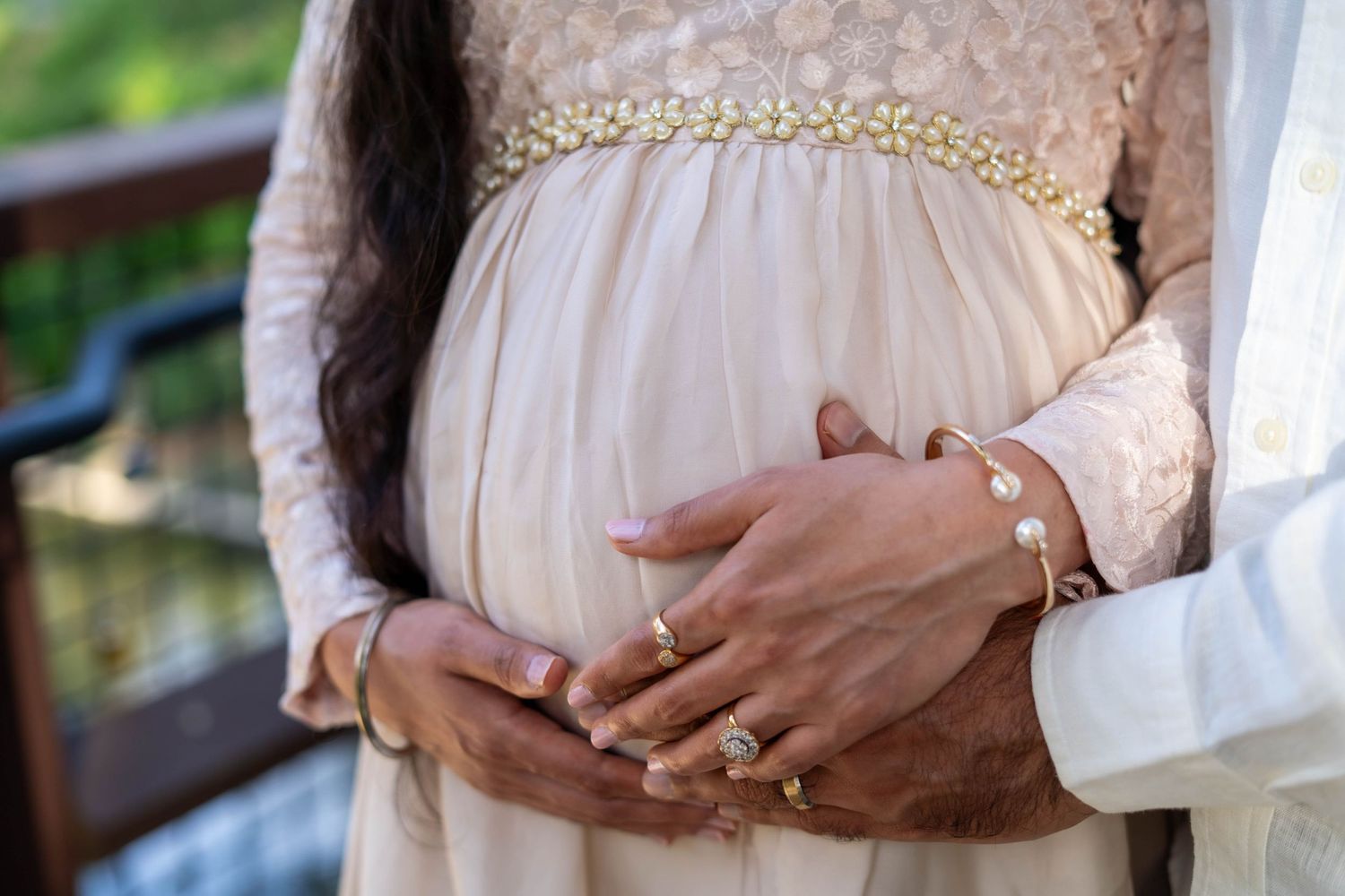 Couple tenderly cradling pregnant belly, wearing elegant gold jewelry and formal attire outdoors.