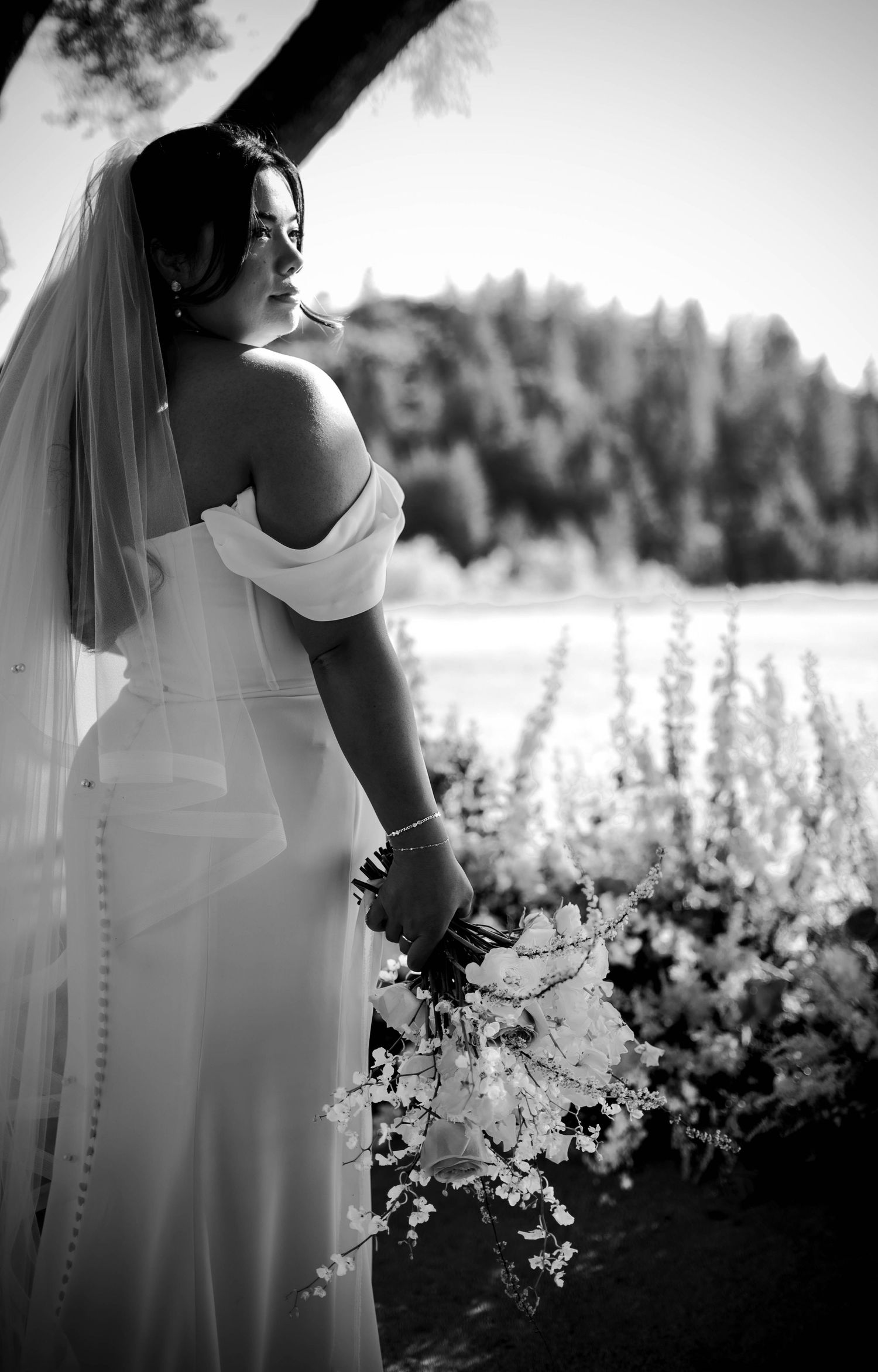 Black and white photo of a bride in an off-shoulder gown and veil holding a bouquet, gazing at a scenic outdoor backdrop.