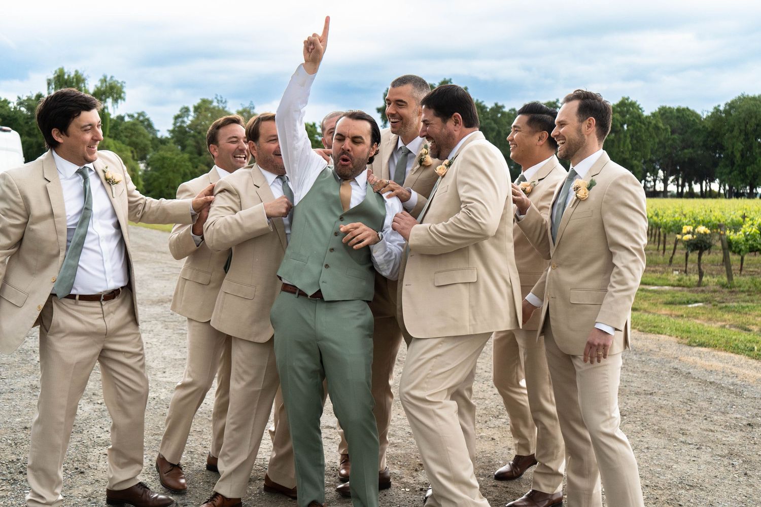 Groomsmen in tan suits pose playfully for group photos at an outdoor wedding venue.