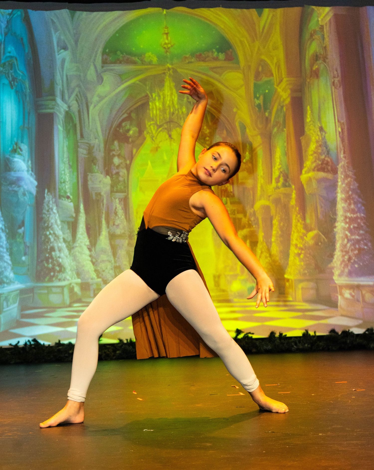 Young ballet dancer performs graceful side stretch on stage with festive Nutcracker backdrop in orange leotard and white tights.