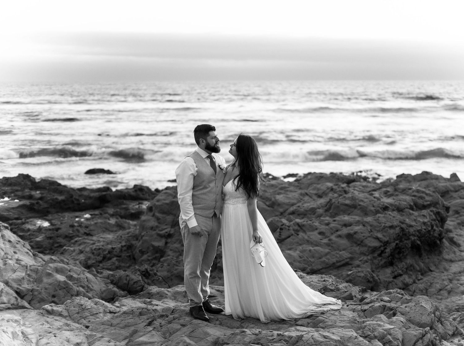 Black and white photo of a couple sharing a kiss on rocky coastal cliffs with ocean waves in the background.