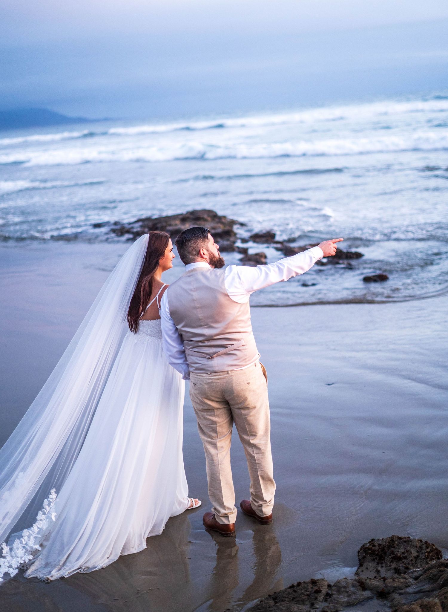 Bride and groom standing on a beach at dusk, groom pointing toward ocean waves, bride wearing white gown and veil.