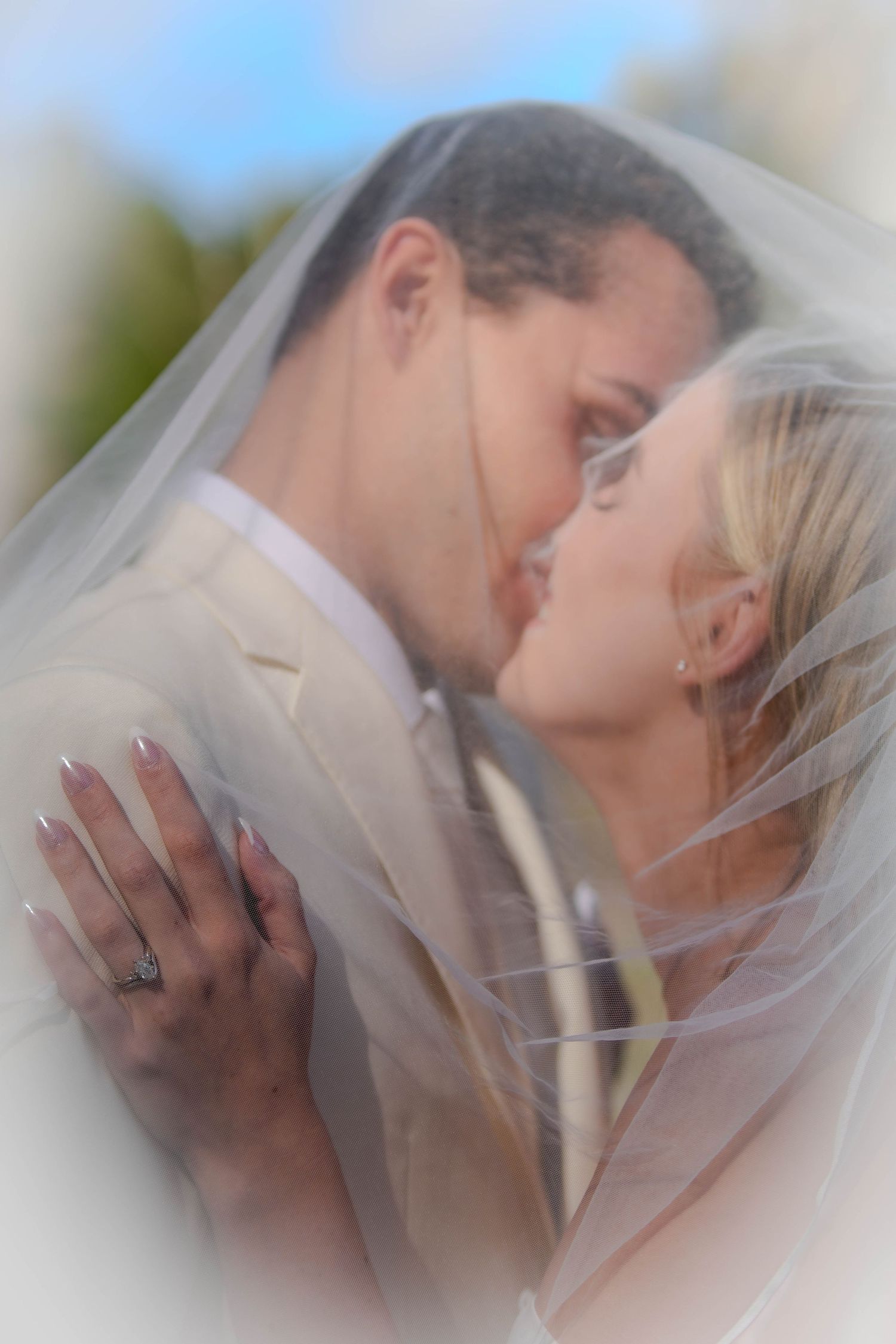 Bride and groom sharing a romantic kiss under a wedding veil outdoors, bride wearing diamond ring.