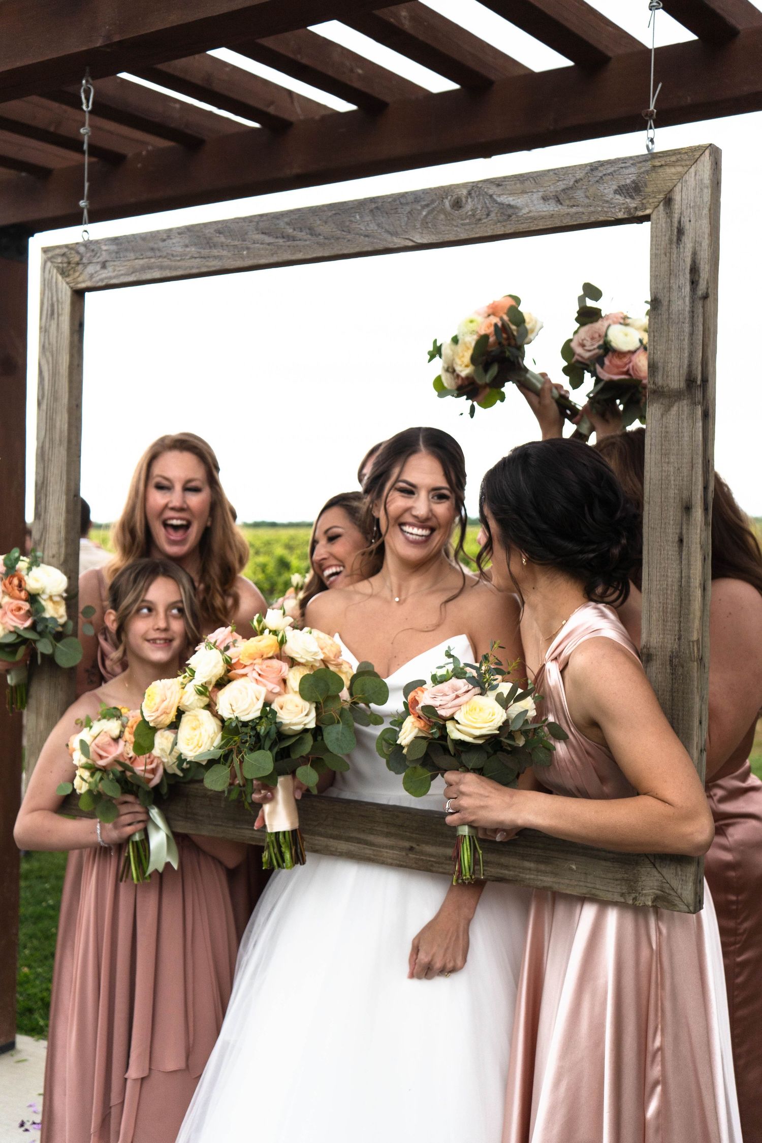 Wedding party members in blush pink dresses and white gown share joyful laughter while holding floral bouquets.