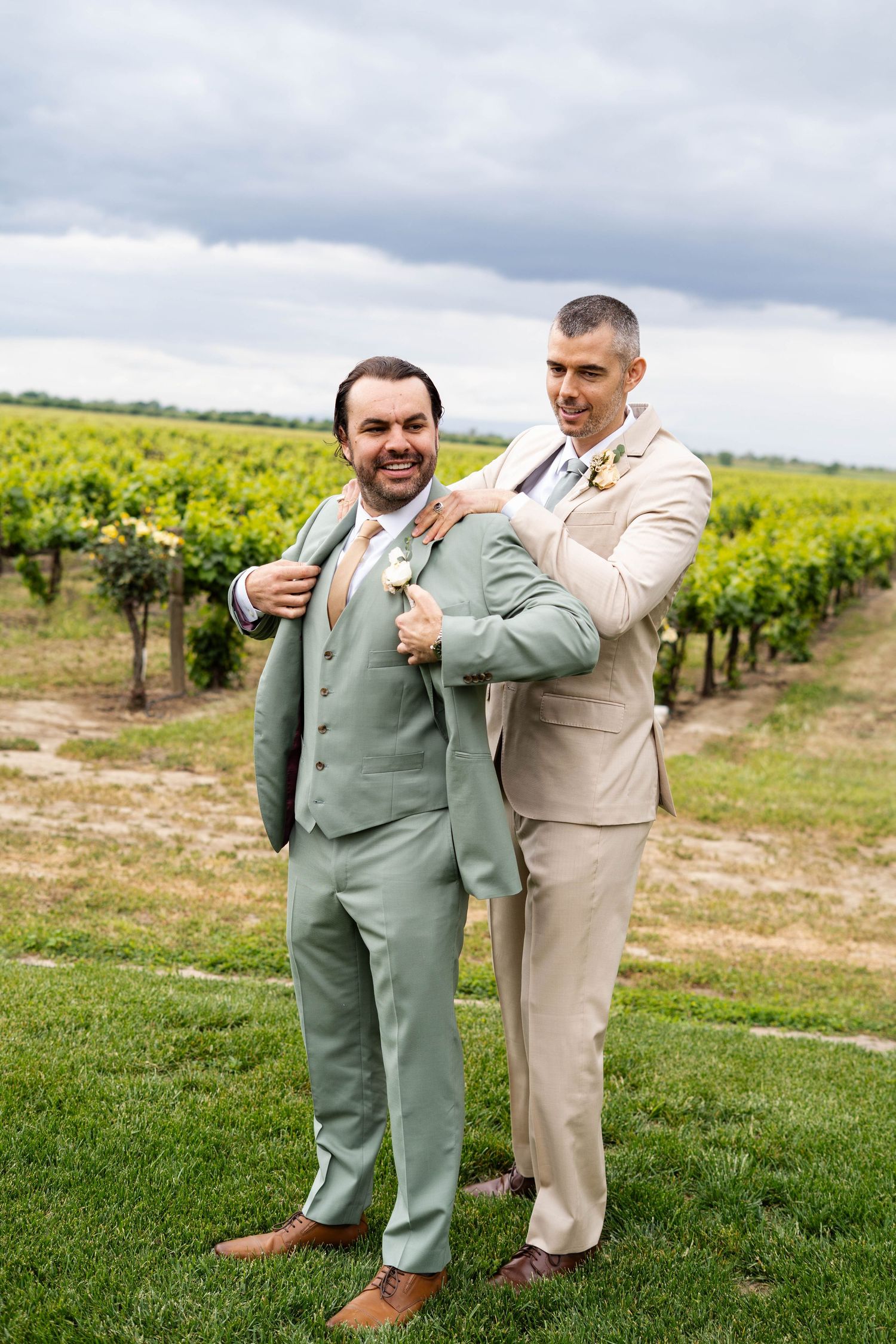 Wedding party members in sage green and tan suits posing in a vineyard setting.