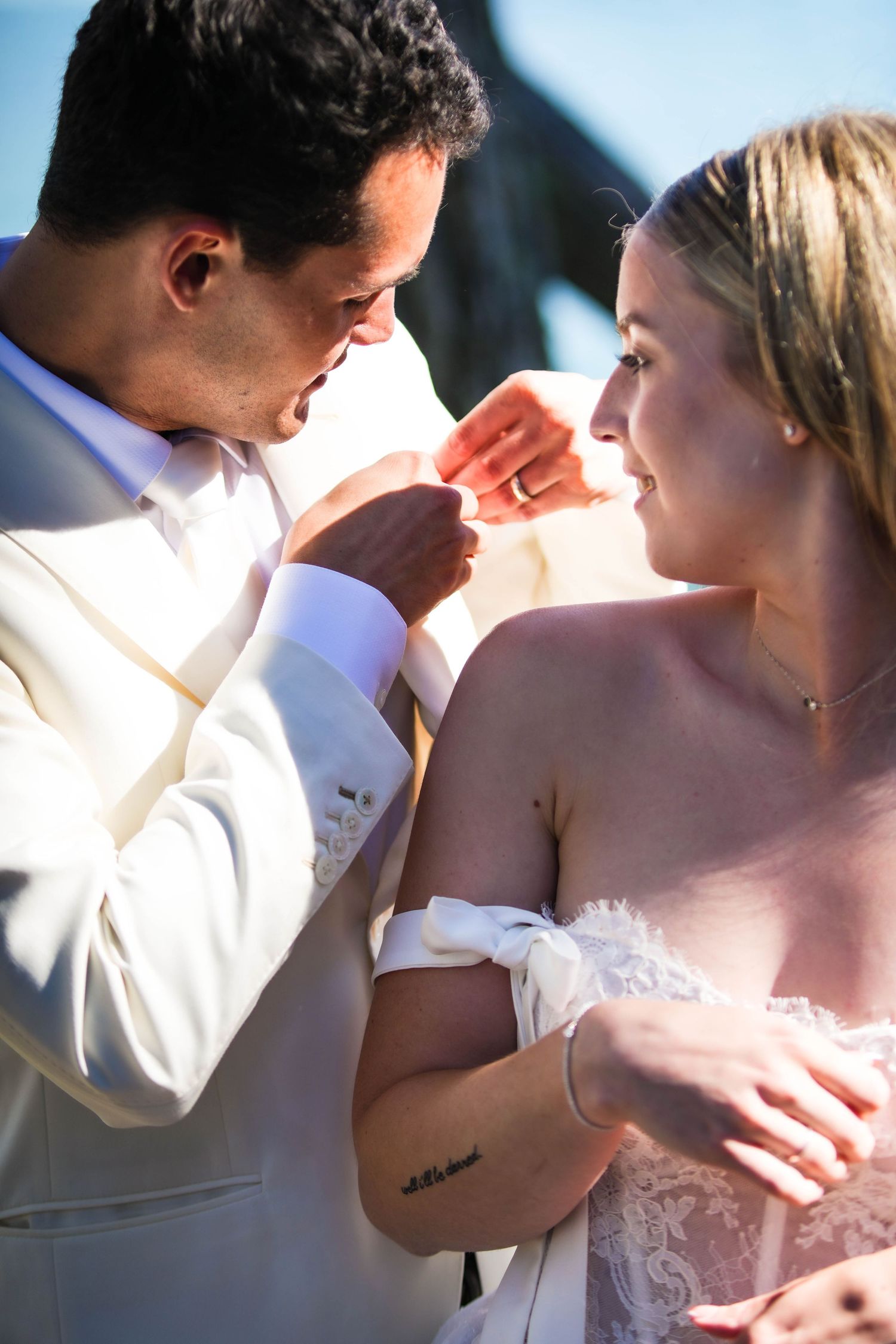 Bride and groom share a tender moment outdoors, groom in white suit kissing her hand, bride in lace dress smiling.