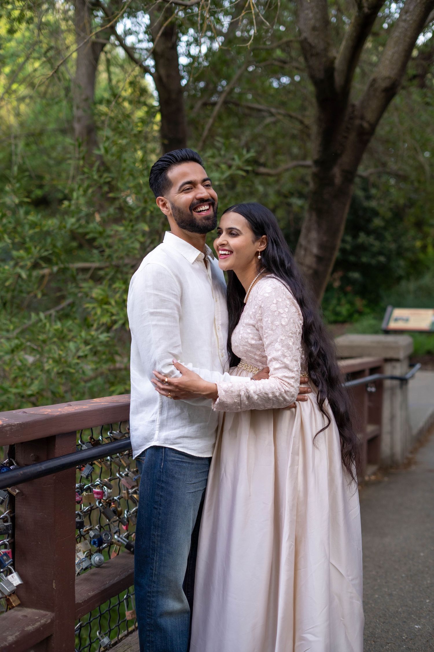 A smiling couple embraces on a bridge surrounded by lush green trees, the woman in a elegant light pink gown.