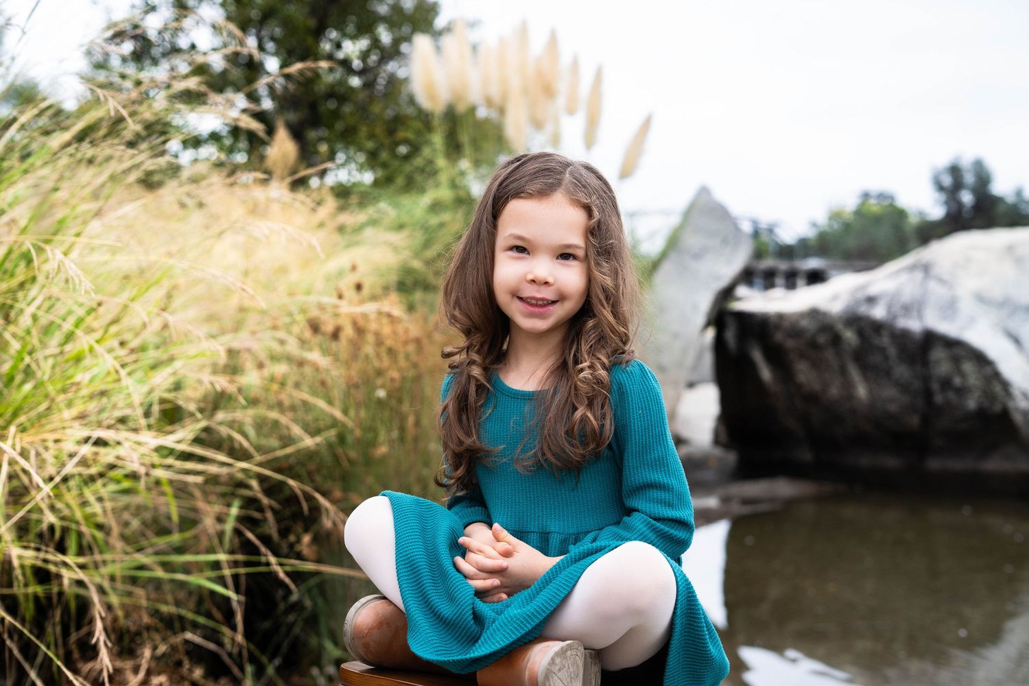 Child in teal sweater poses playfully outdoors among tall grass and rocks.