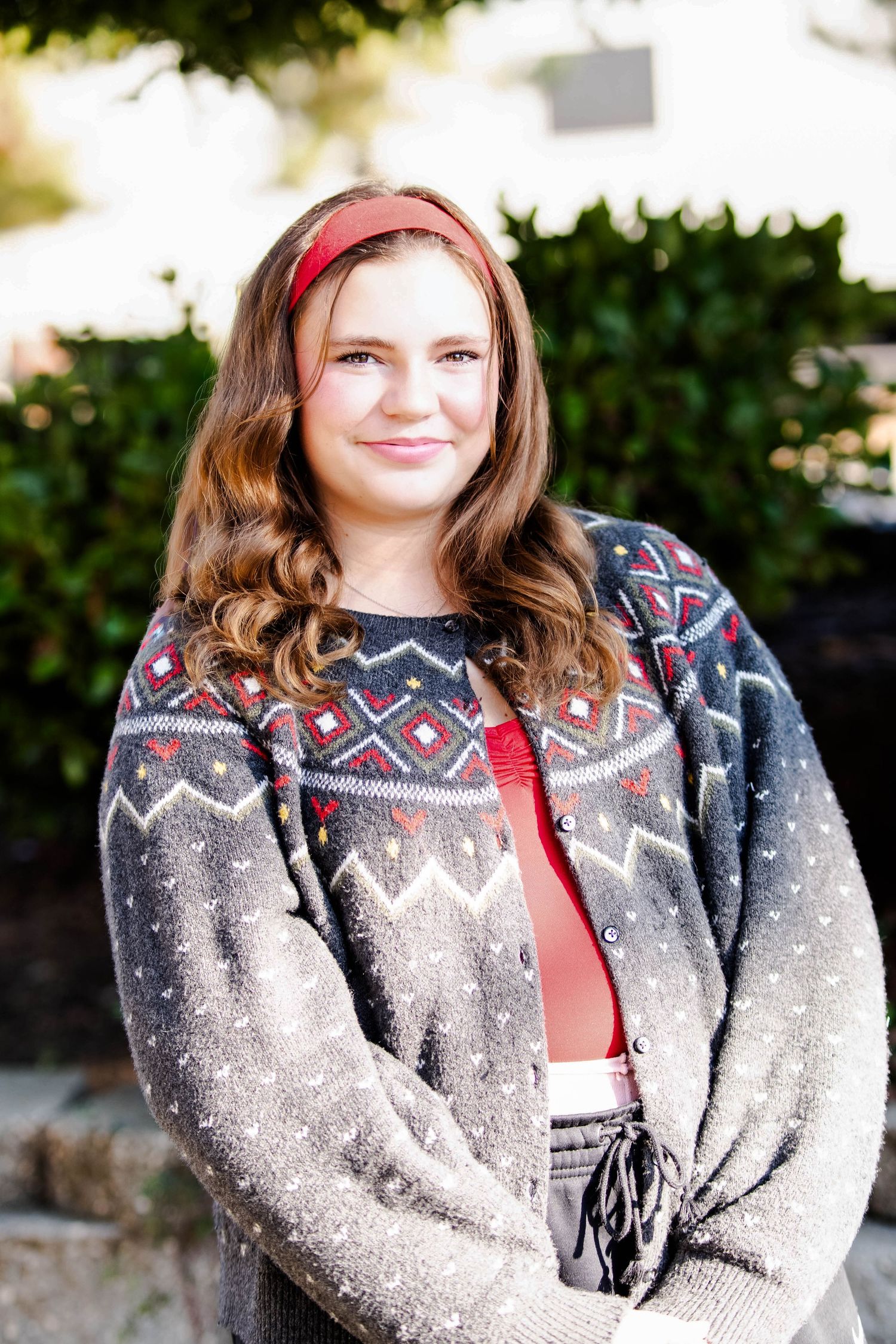 Young woman in a Fair Isle cardigan and red headband smiles outdoors near green hedges.