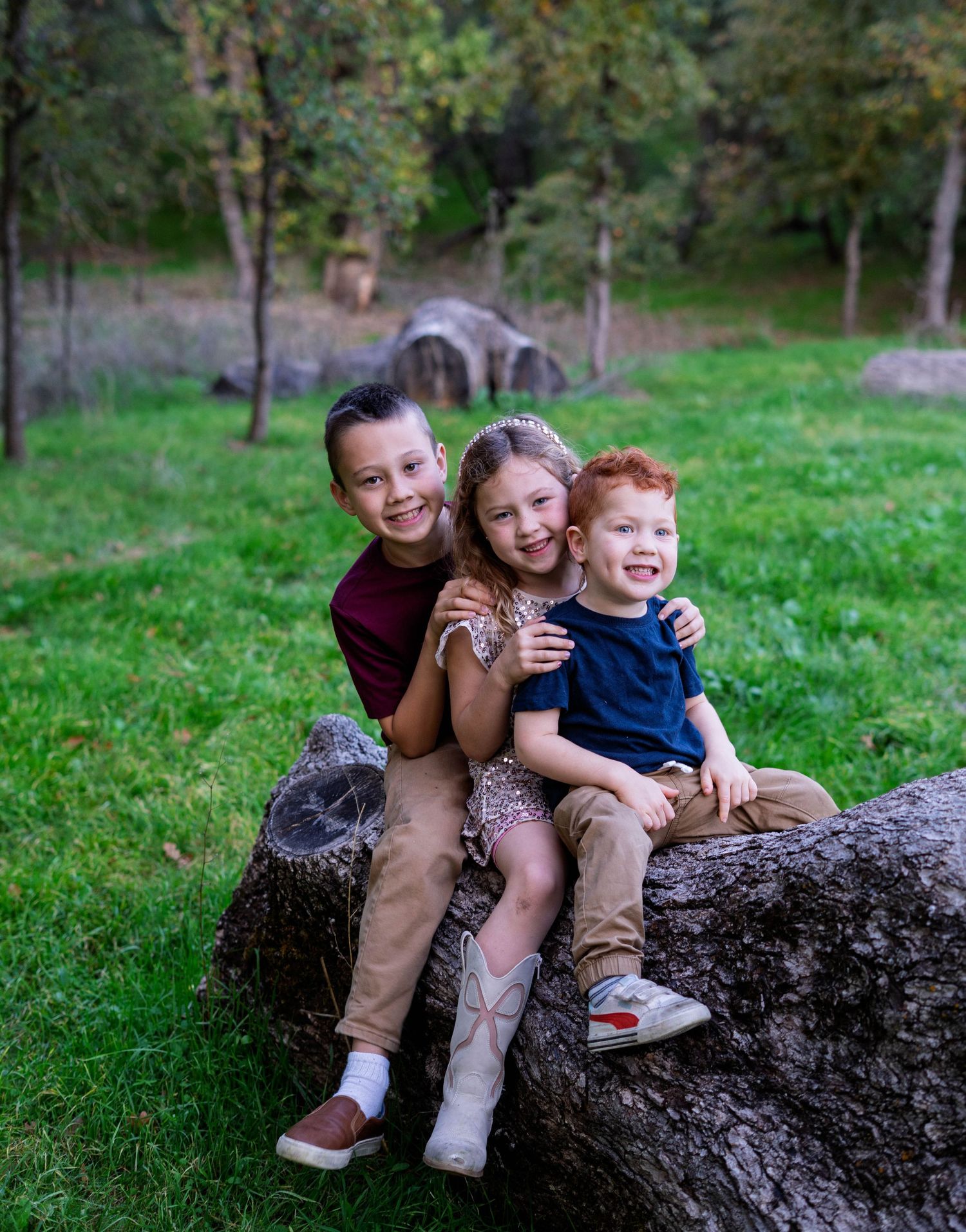 Three children sit together and laugh while perched on a fallen tree trunk in a grassy outdoor setting.