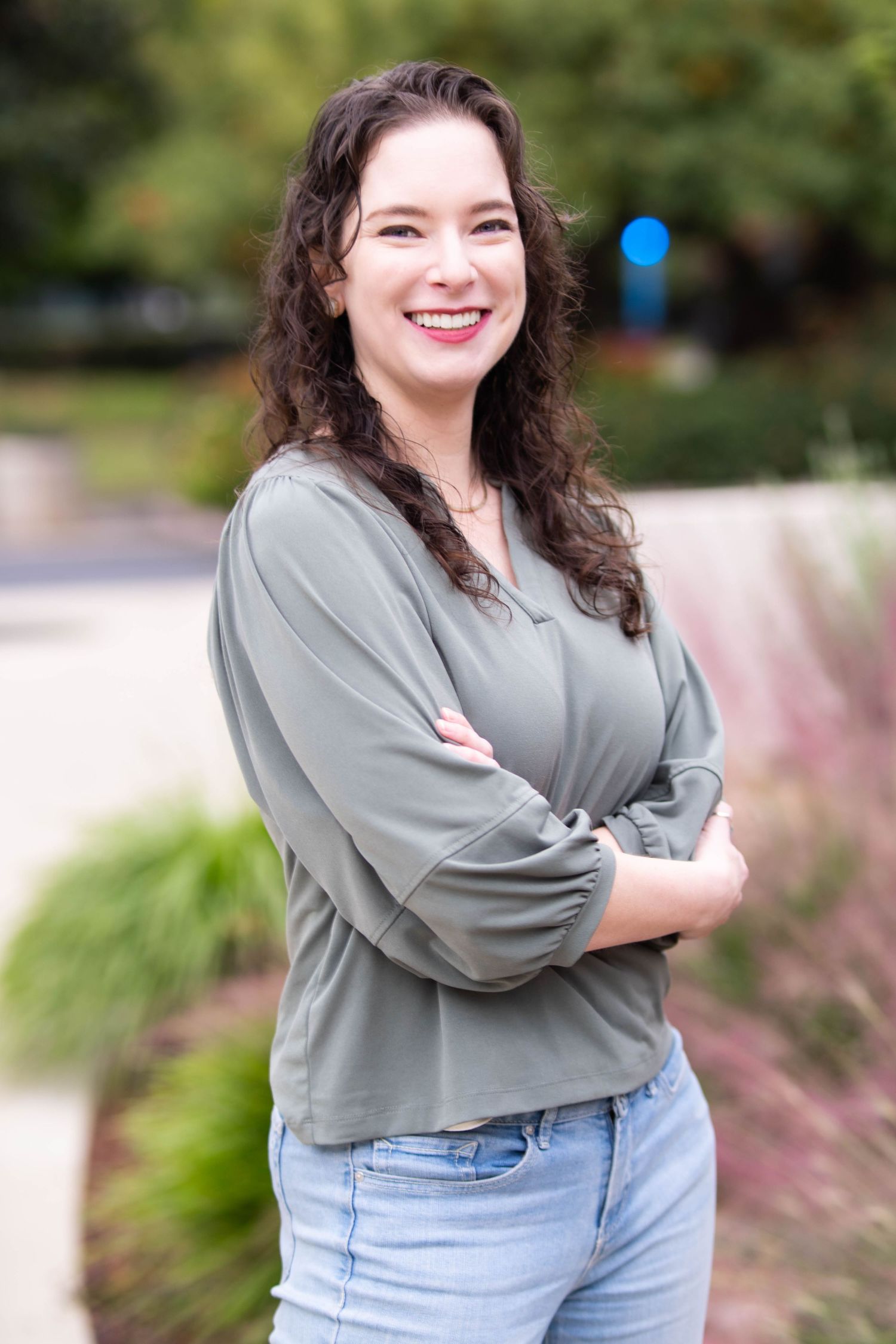 A person in a gray sweater and light jeans poses outdoors against a natural garden background.