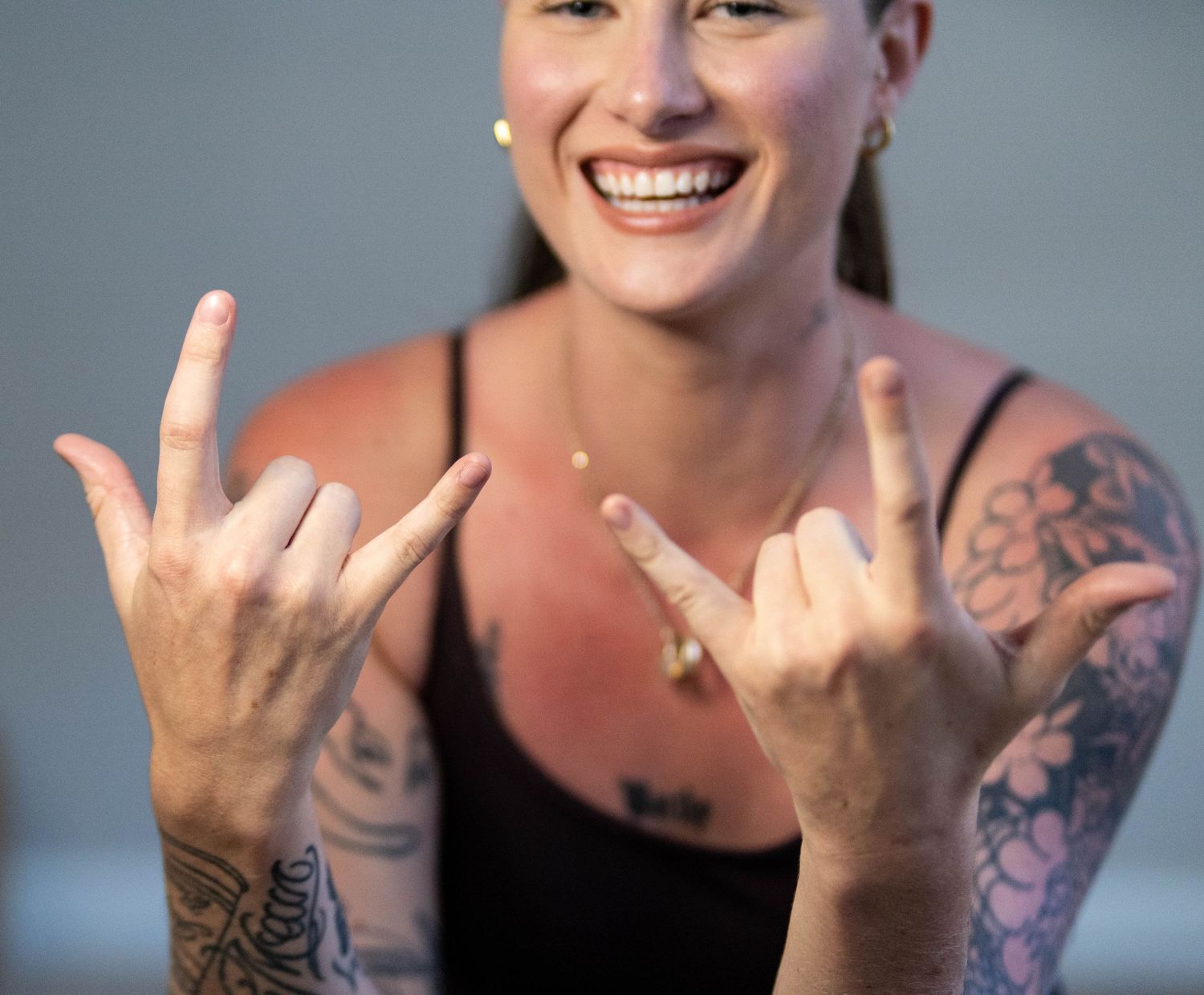 Person wearing black tank top showing rock and roll hand signs with visible arm tattoos.