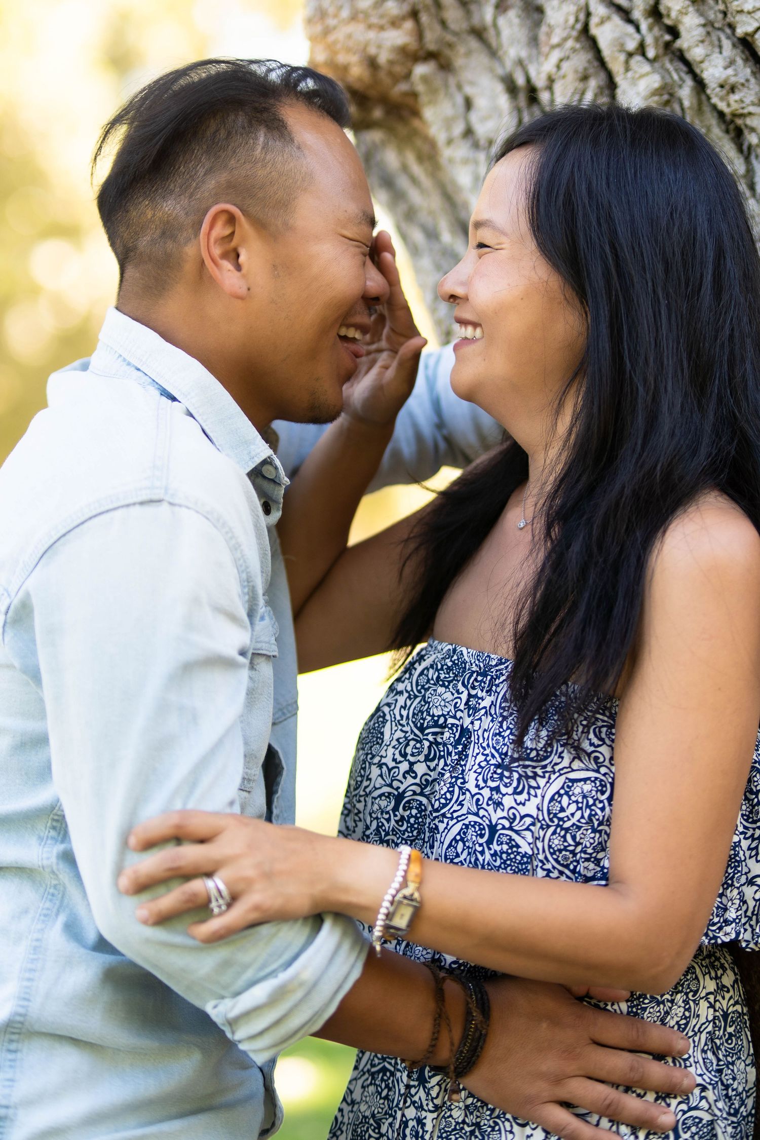 A couple shares a joyful laugh while embracing outdoors near a tree trunk.