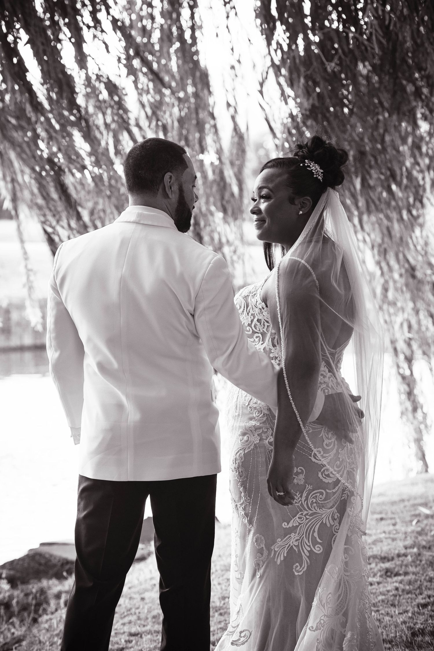 Black and white portrait of newlyweds walking beneath dramatic willow tree branches.