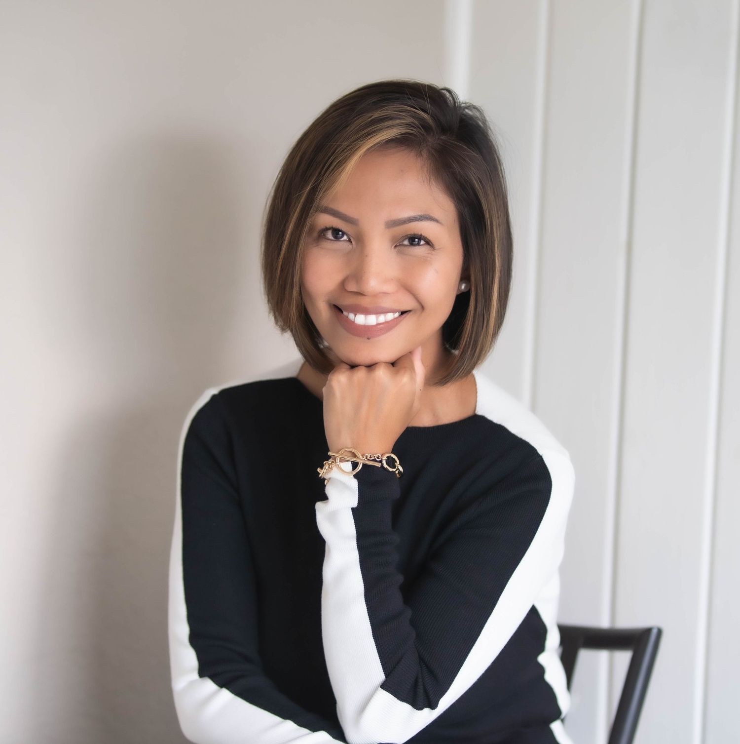 A person in a black and white sweater poses with hand under chin against white wall.