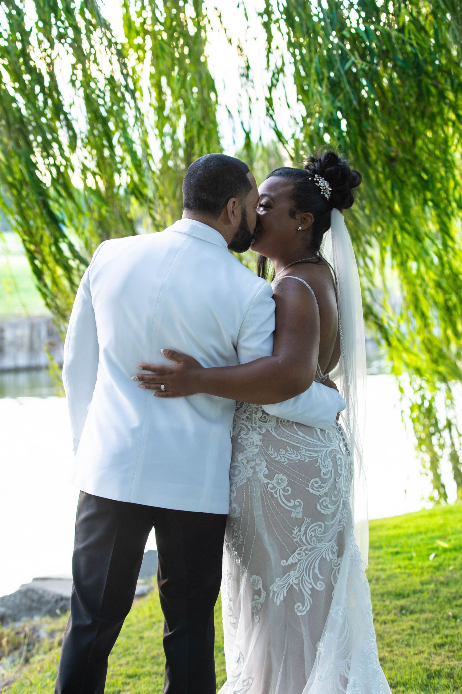 Newlyweds sharing an intimate moment under weeping willow trees by a waterfront.