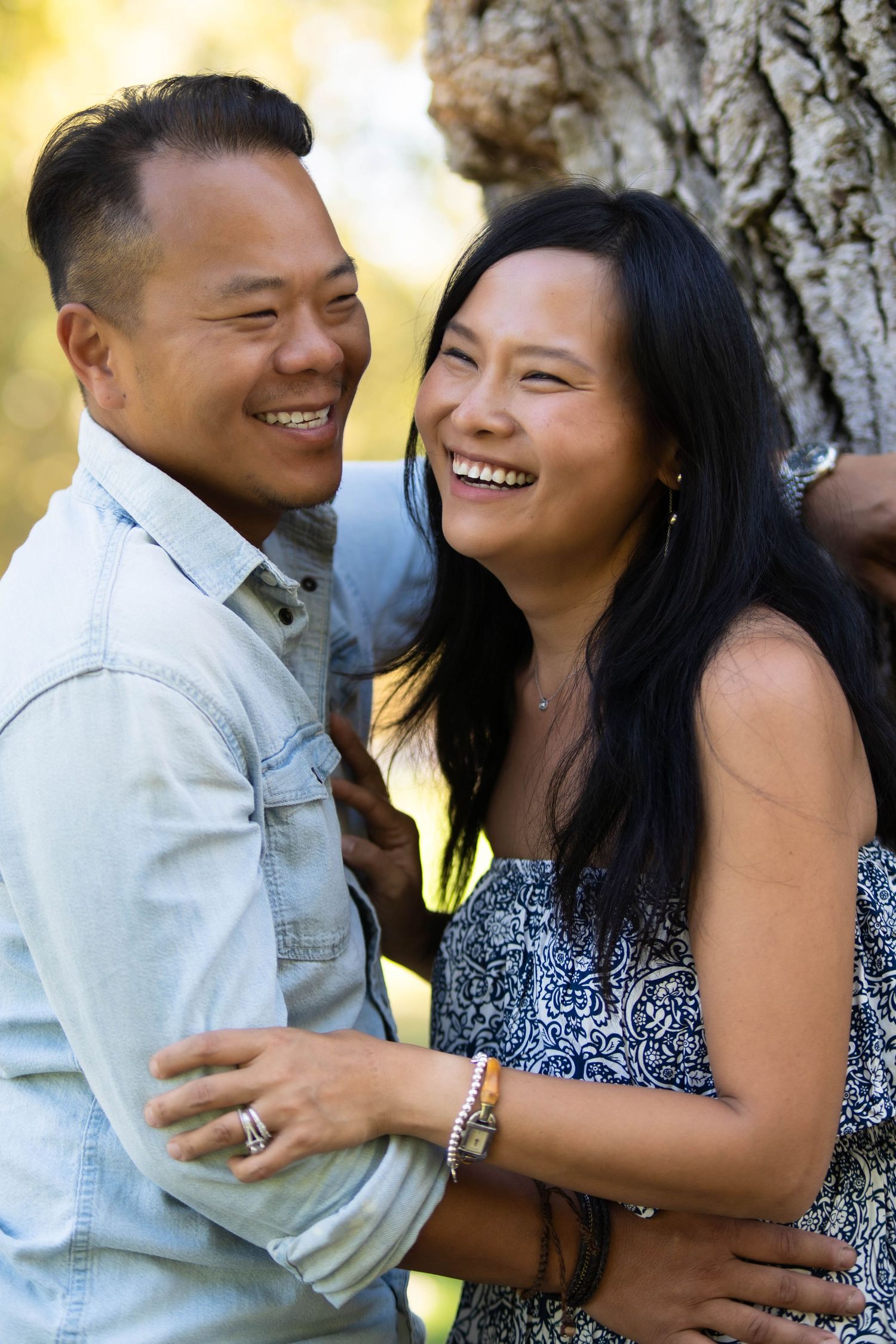 A couple shares genuine laughter while embracing against a tree in natural lighting.