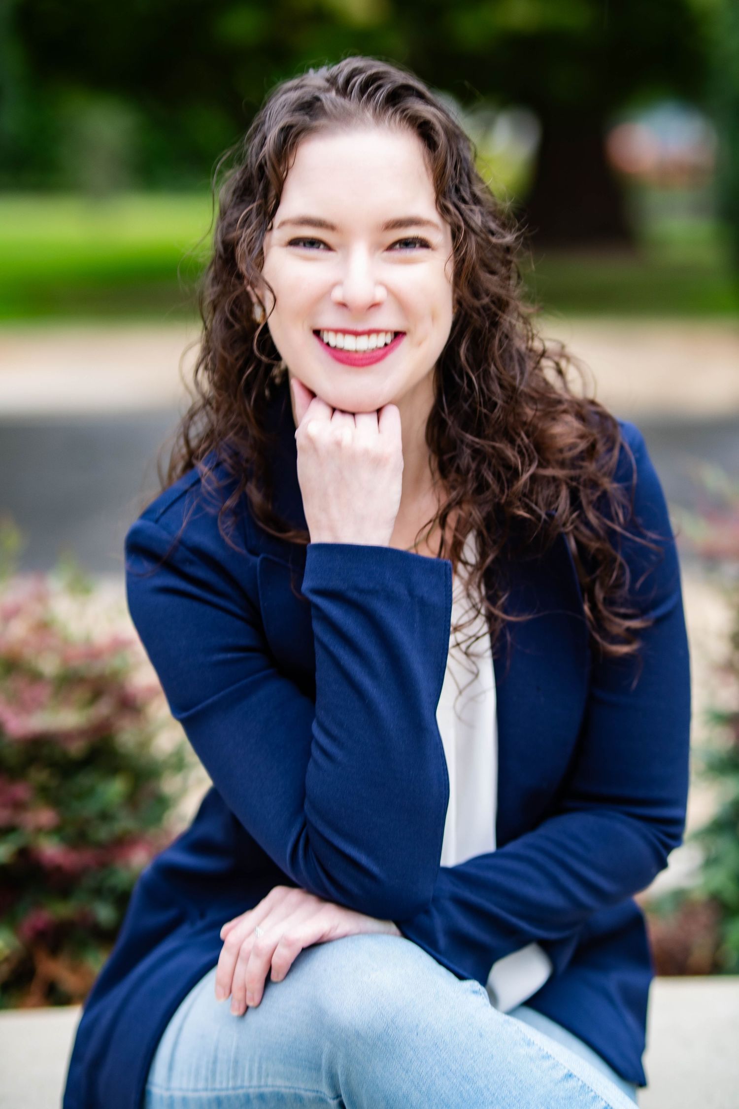 A person in a navy cardigan and light jeans sits outdoors smiling with curly brown hair and natural makeup.
