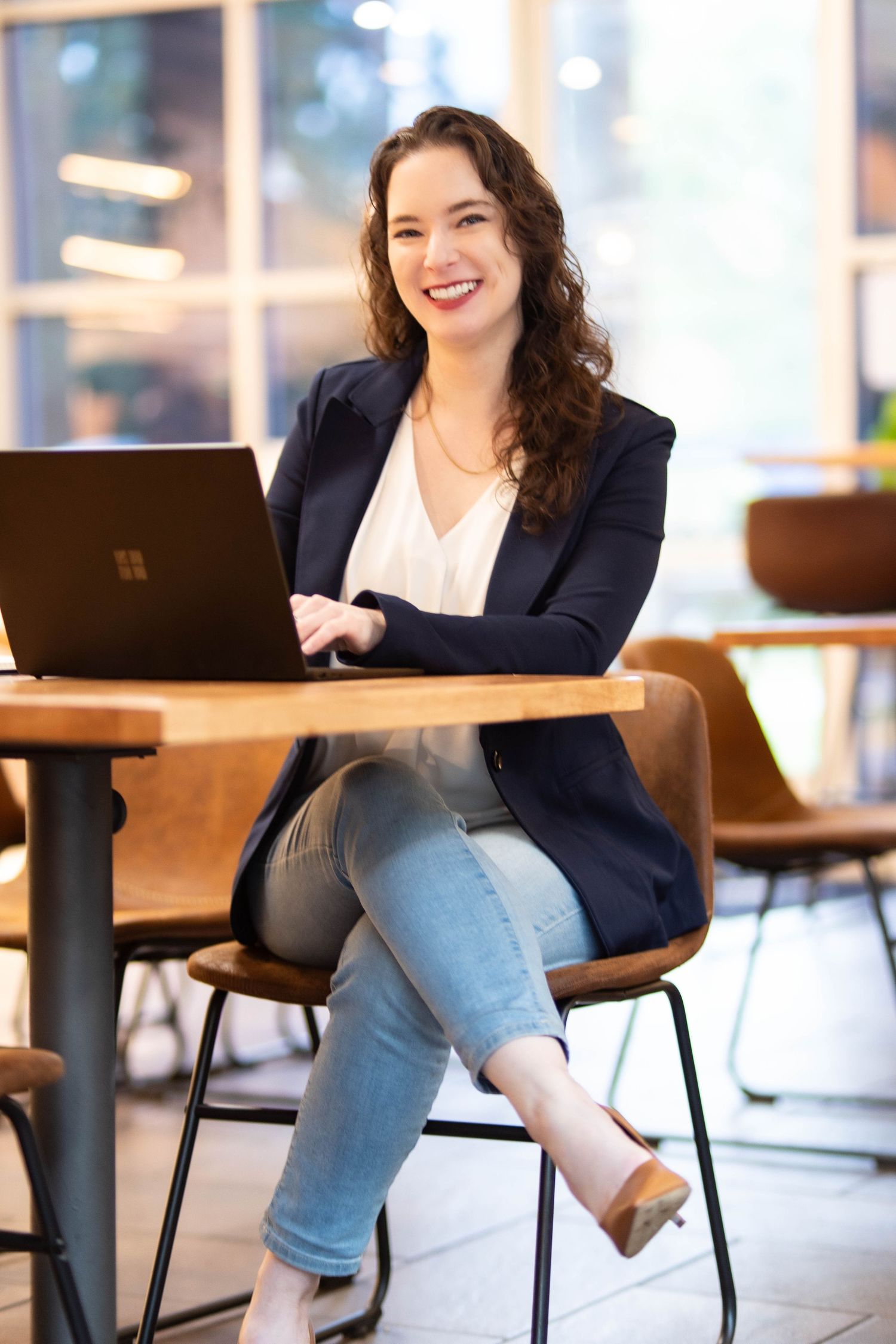 Professional wearing navy blazer and jeans sits casually at wooden desk in modern office setting.