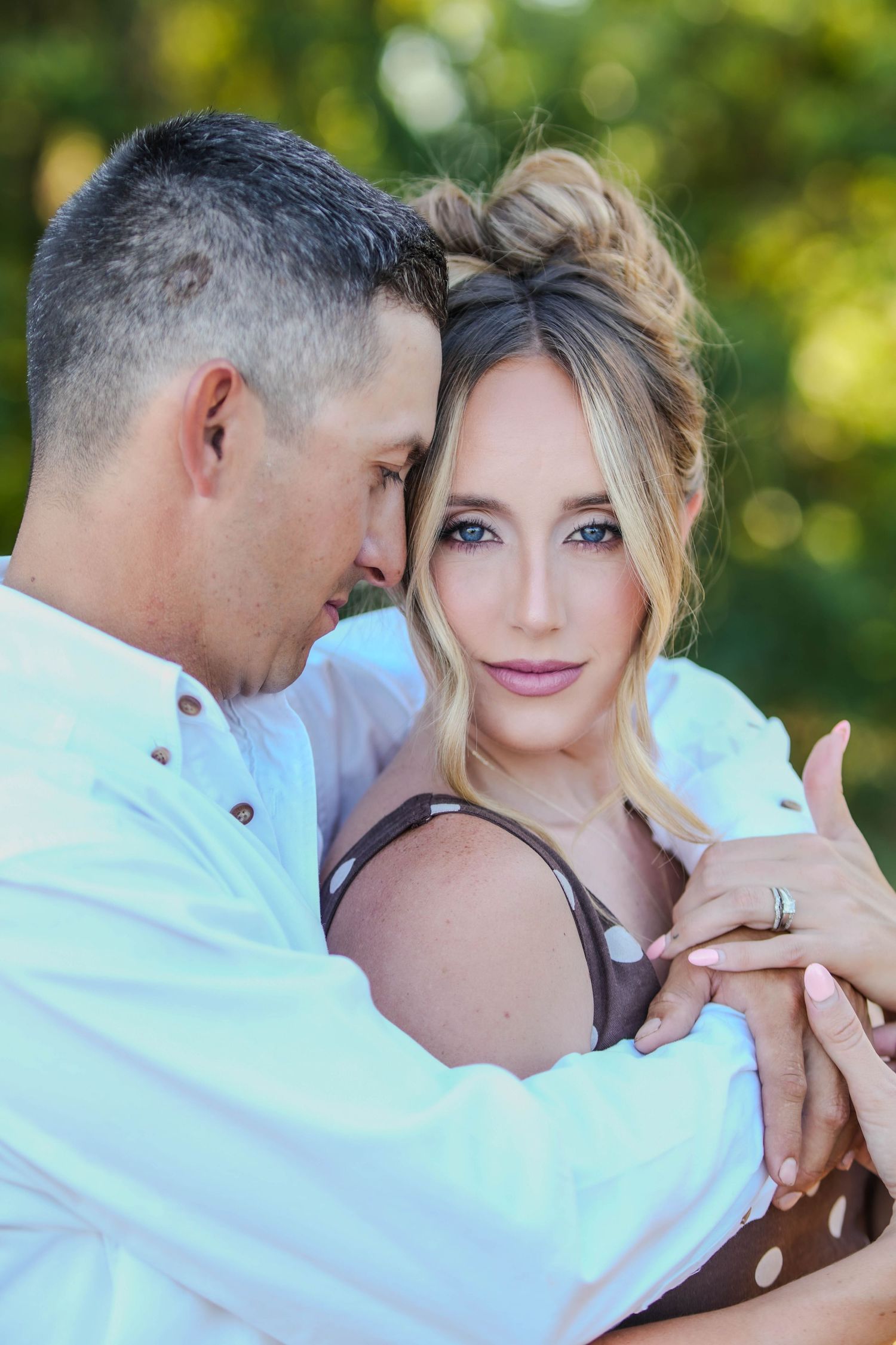 A couple embraces tenderly outdoors, the woman gazing at the camera while the man holds her close in a romantic portrait.