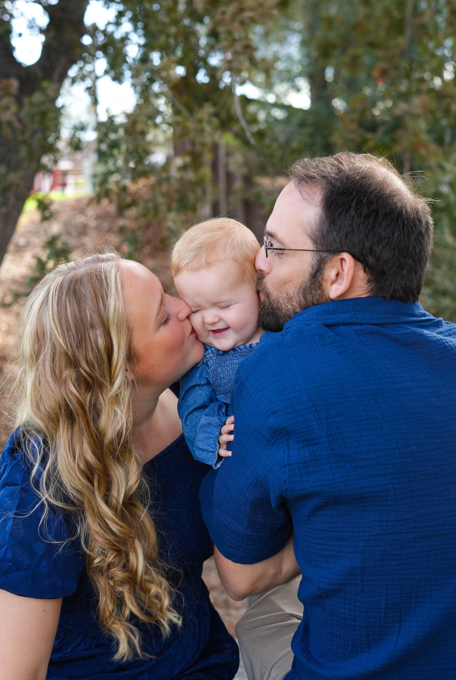 A happy family in blue outfits, parents kissing their smiling red-haired baby outdoors among green trees.