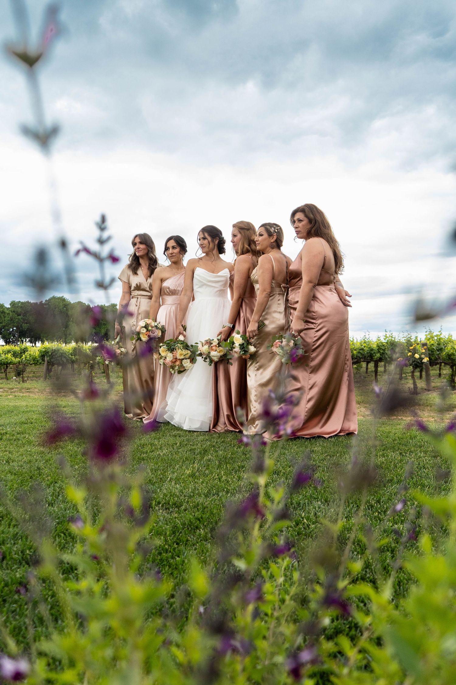 Bridal party in blush dresses posed together in a vineyard with purple wildflowers.